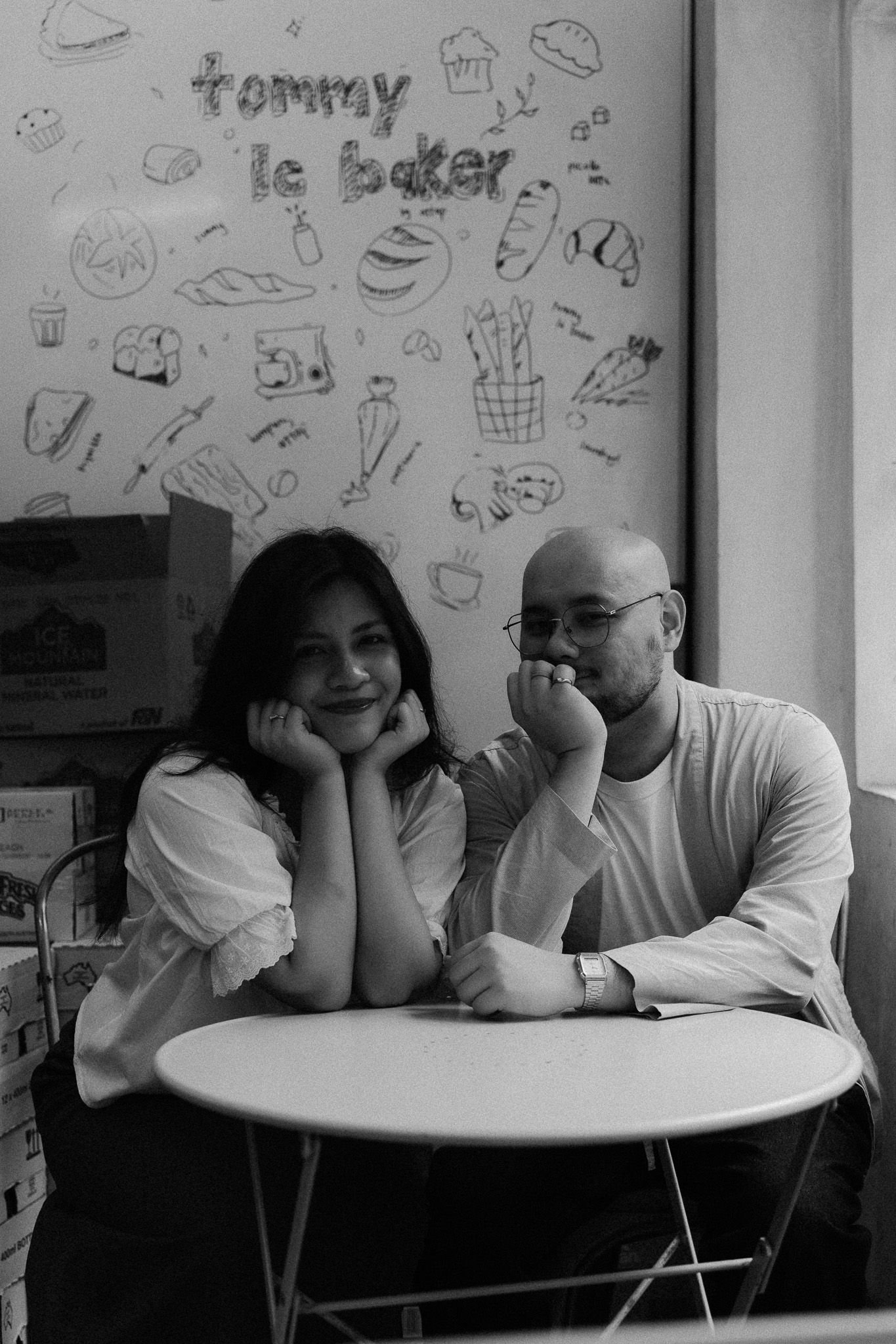 Black and white photo of a couple sitting at a round table in front of a Tommy Le Baker wall mural.