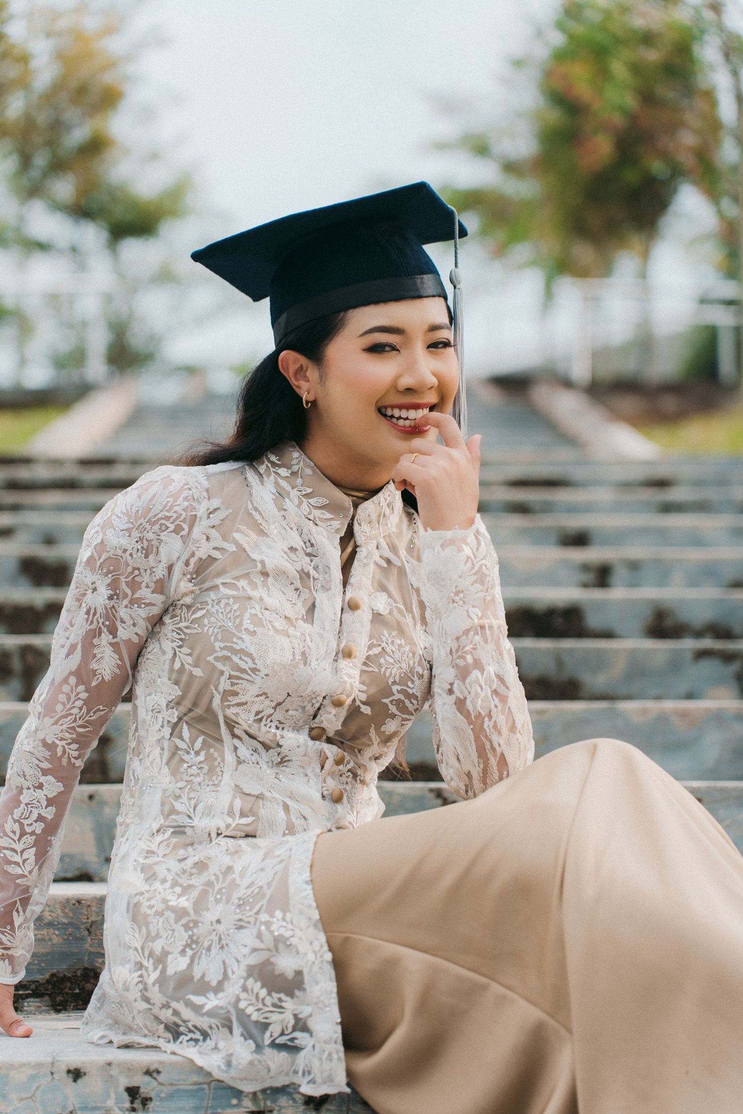 Graduate in white lace top and graduation cap sits on bleachers during outdoor photoshoot.