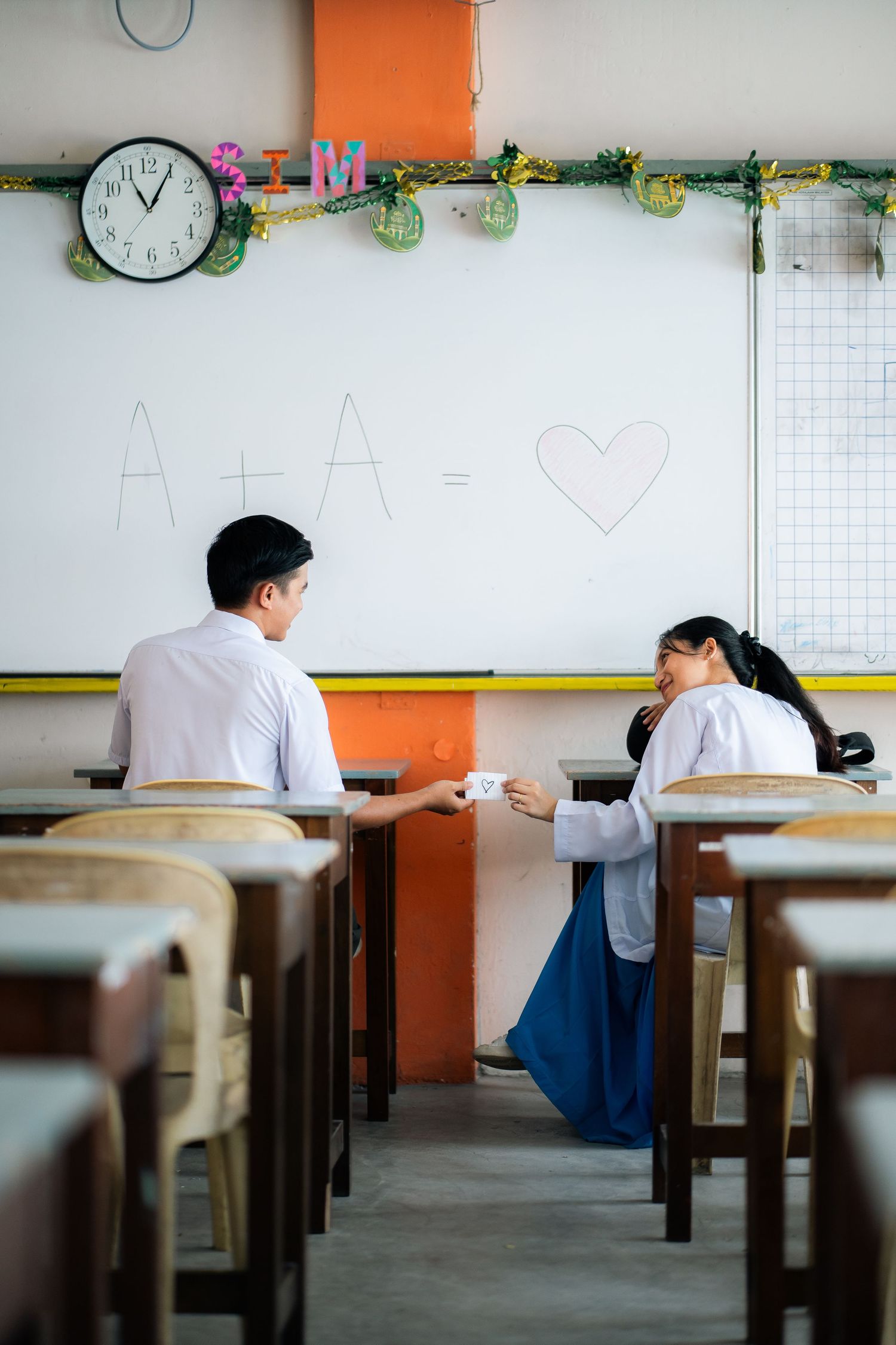 Two students sit at desks in a classroom with an orange accent wall and wall clock visible.