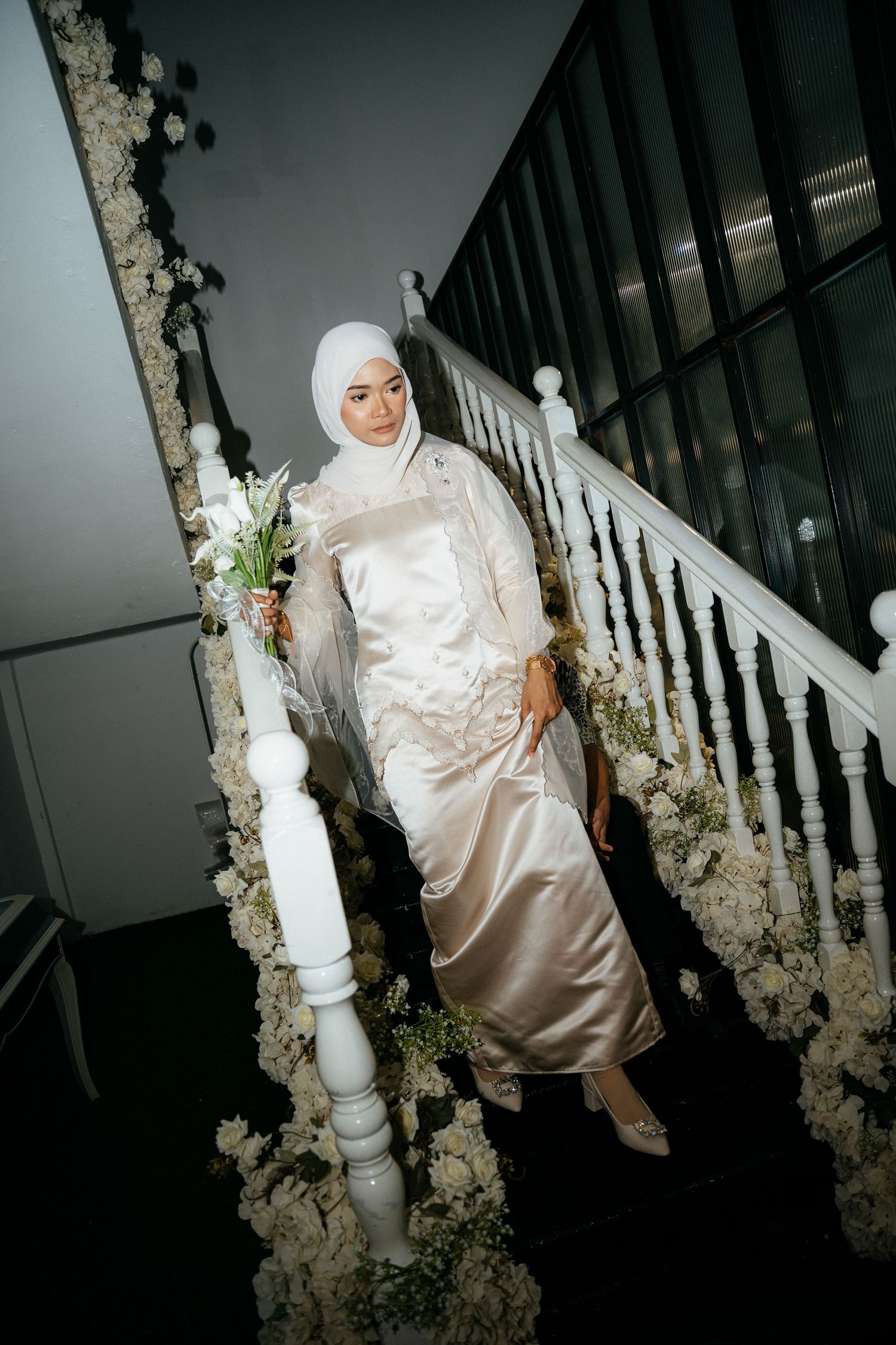 Person in white satin dress and hijab poses on an elegant staircase decorated with white floral arrangements.