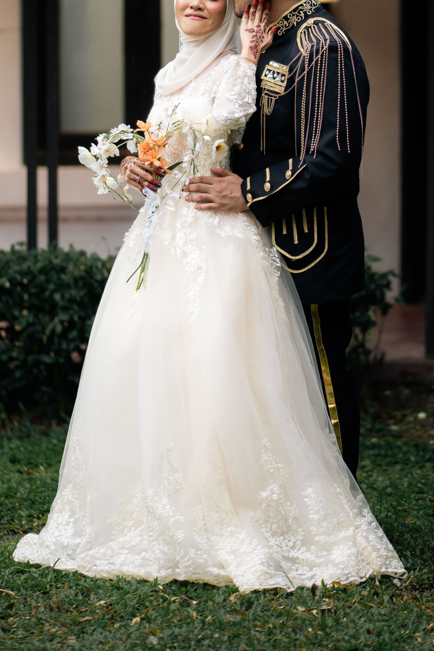 Muslim bride in white lace wedding gown and hijab stands with groom in military uniform holding a bouquet.