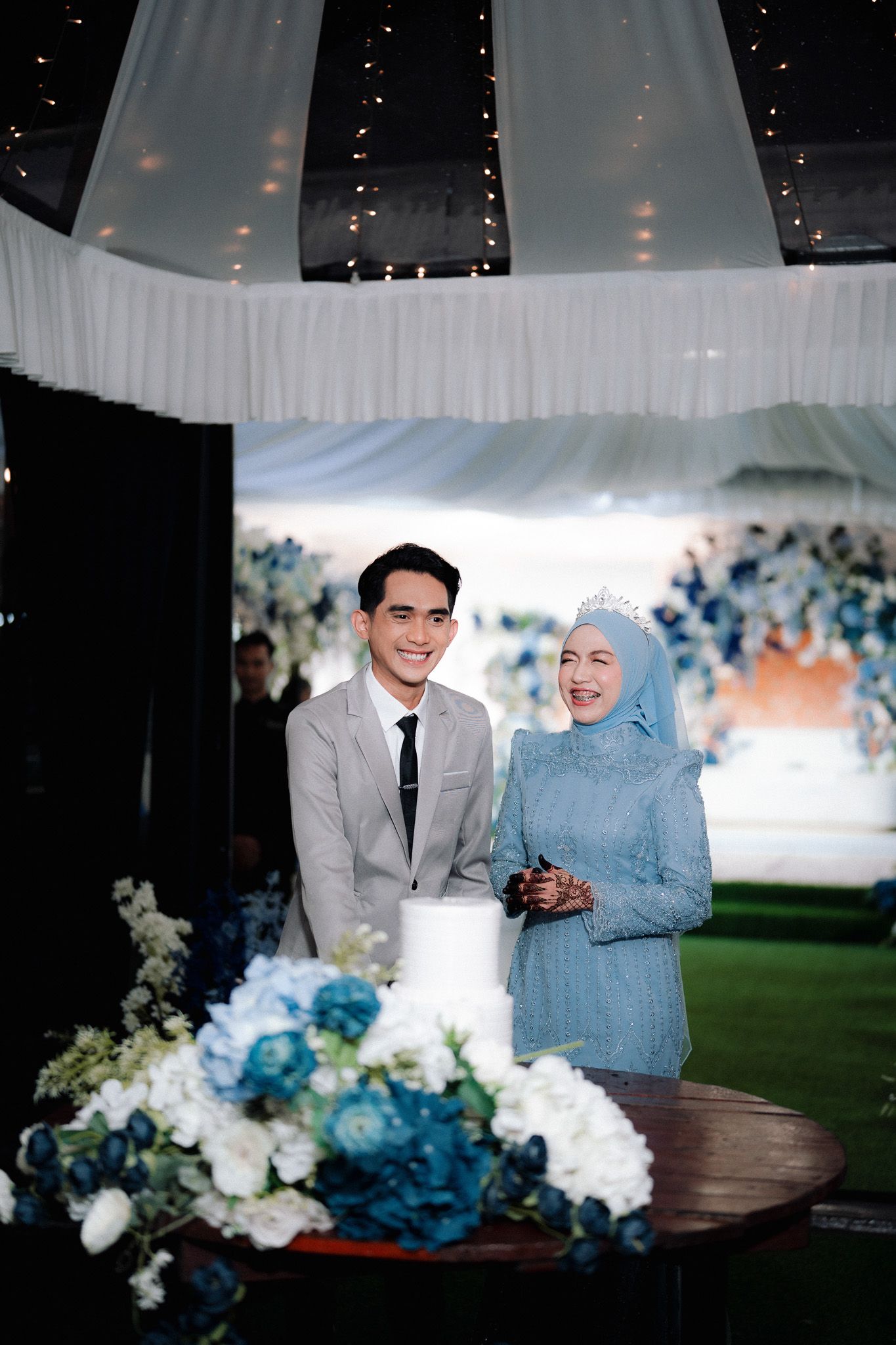 Muslim couple smiling at wedding cake table with blue floral arrangements under elegant draped tent.