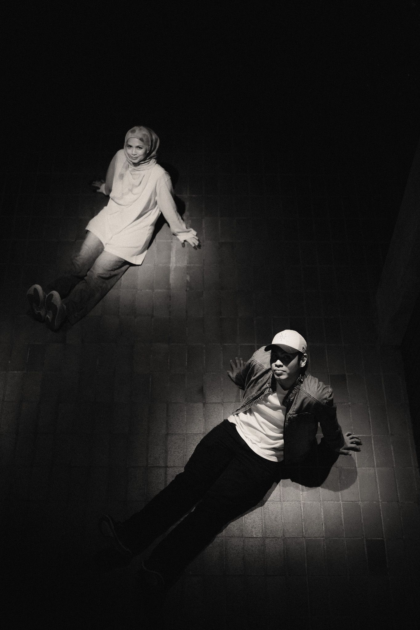 Black and white photo of two people lying on a tiled floor under dramatic spotlight lighting.
