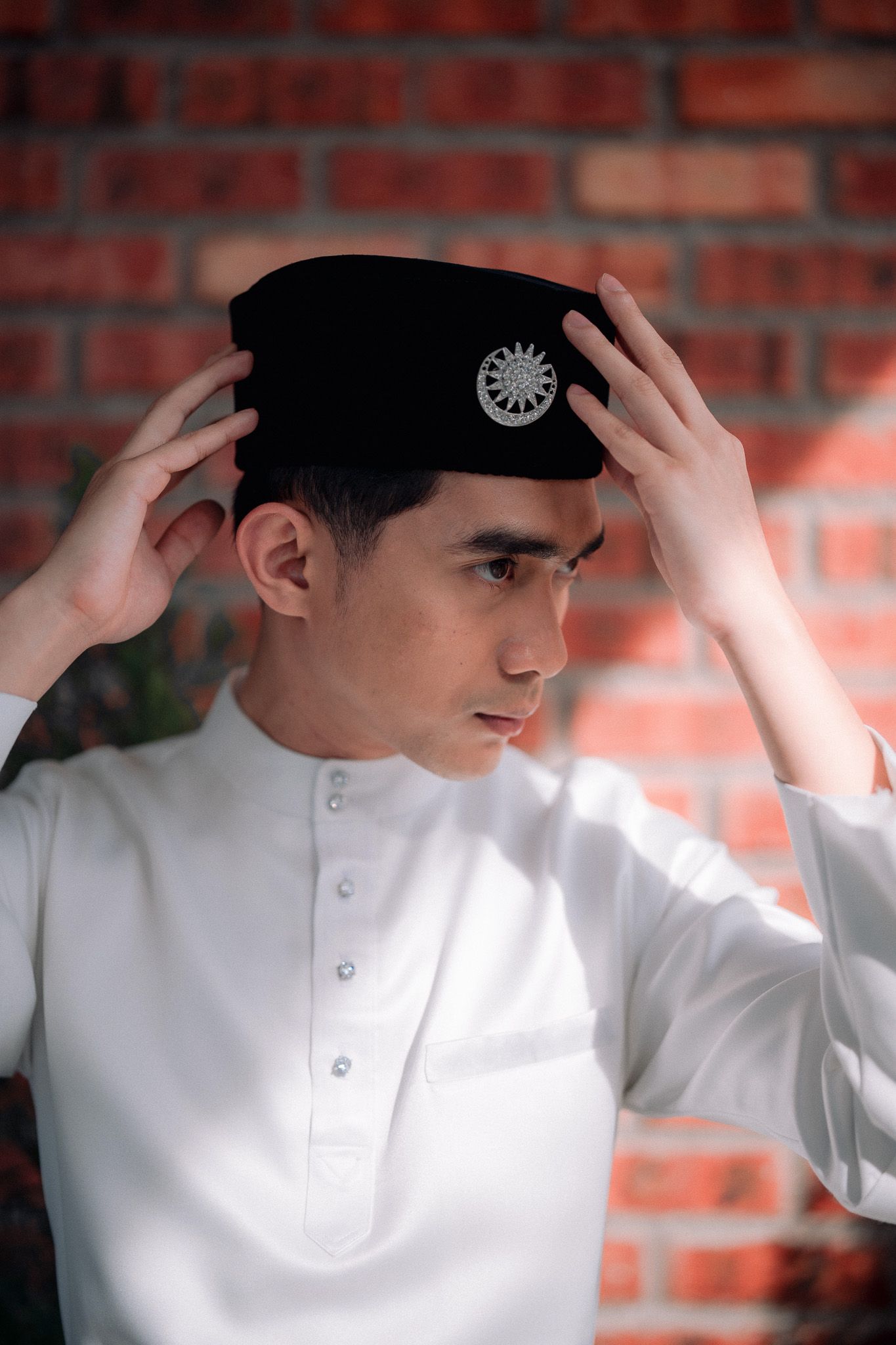 Young Muslim man in white baju melayu adjusting black songkok cap against brick wall background.