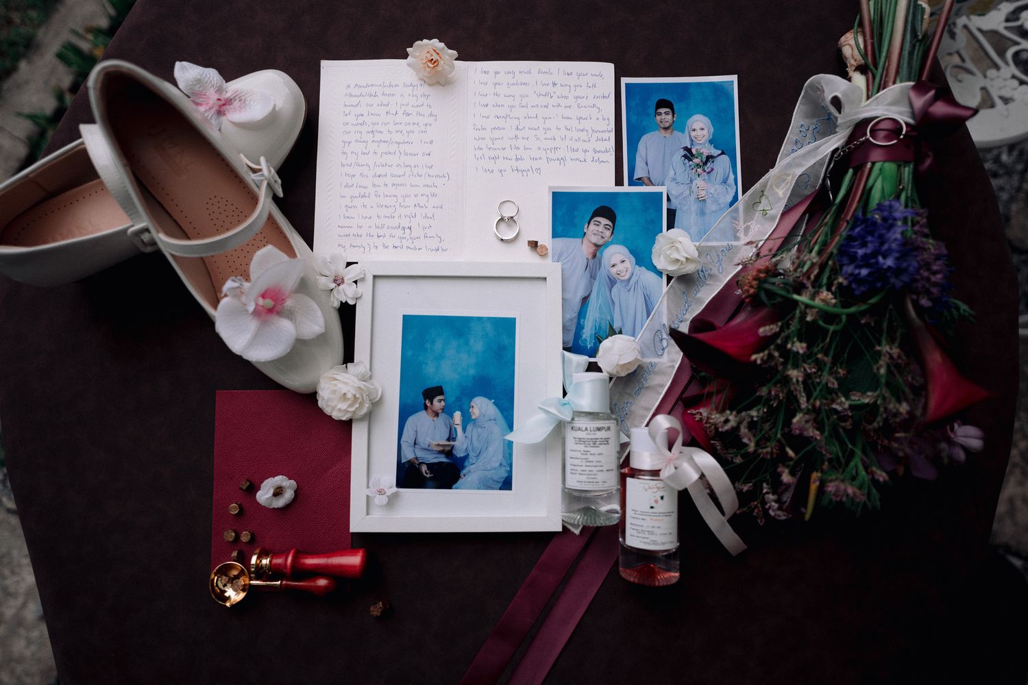 Wedding flat lay with bridal shoes, photos, handwritten letter, flowers, and accessories arranged on a dark surface.
