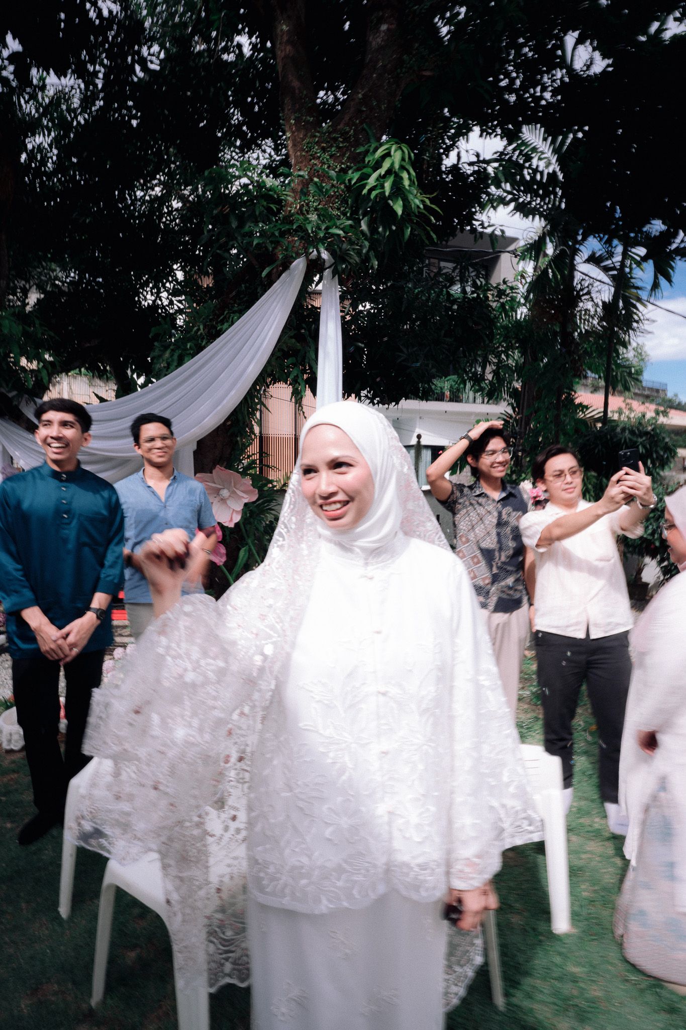 Smiling bride in white hijab and lace dress at outdoor wedding ceremony with guests celebrating in background.