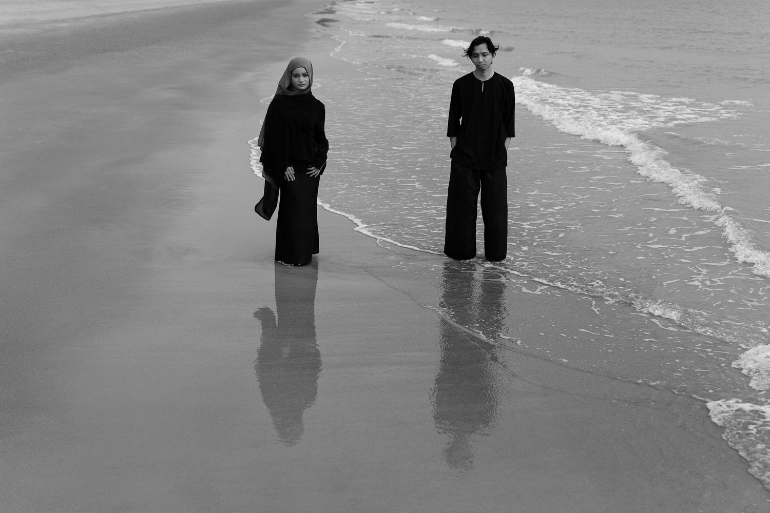 Two people in black clothing stand on a wet beach shore in a dramatic black and white photograph.