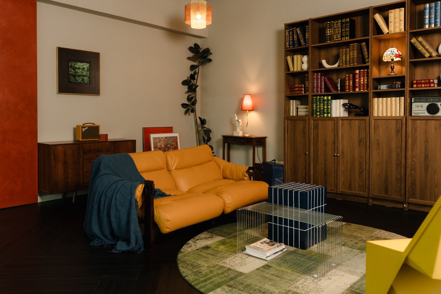 Cozy living room with orange leather sofa, blue throw, wooden bookshelf filled with books, and a glass coffee table on green rug.
