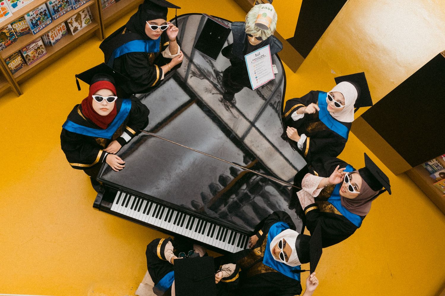 Graduates in academic dress positioned around a grand piano in artistic overhead shot.