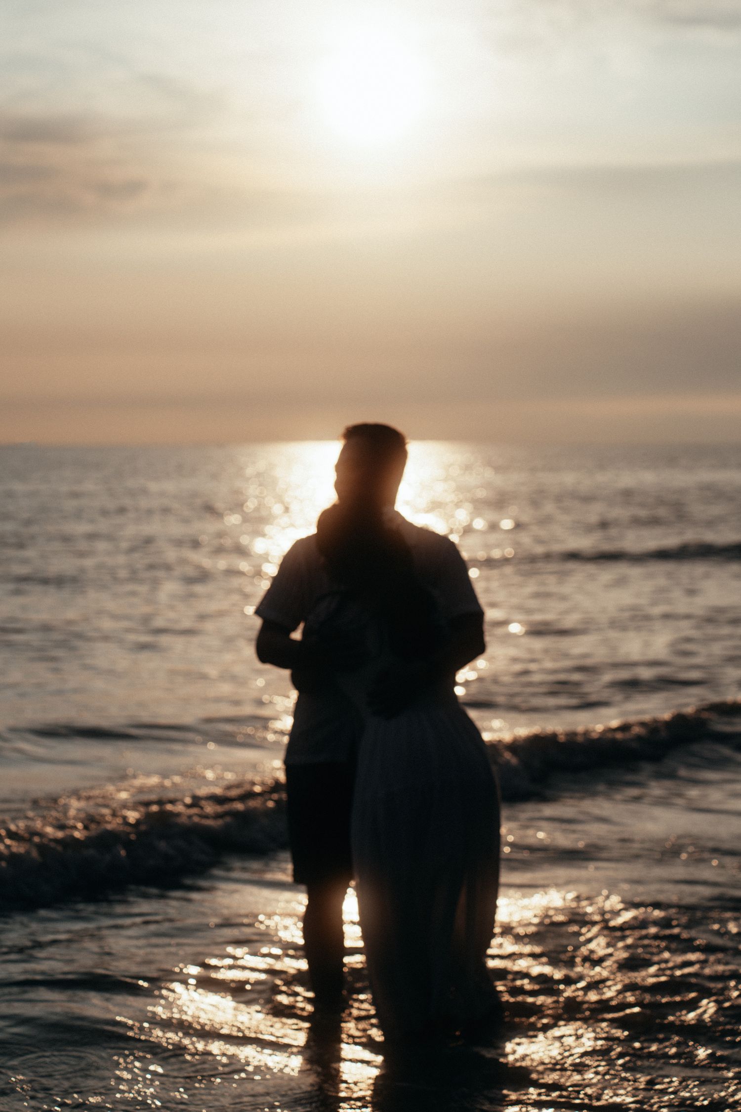Silhouetted couple stands together in shallow ocean water during golden hour.