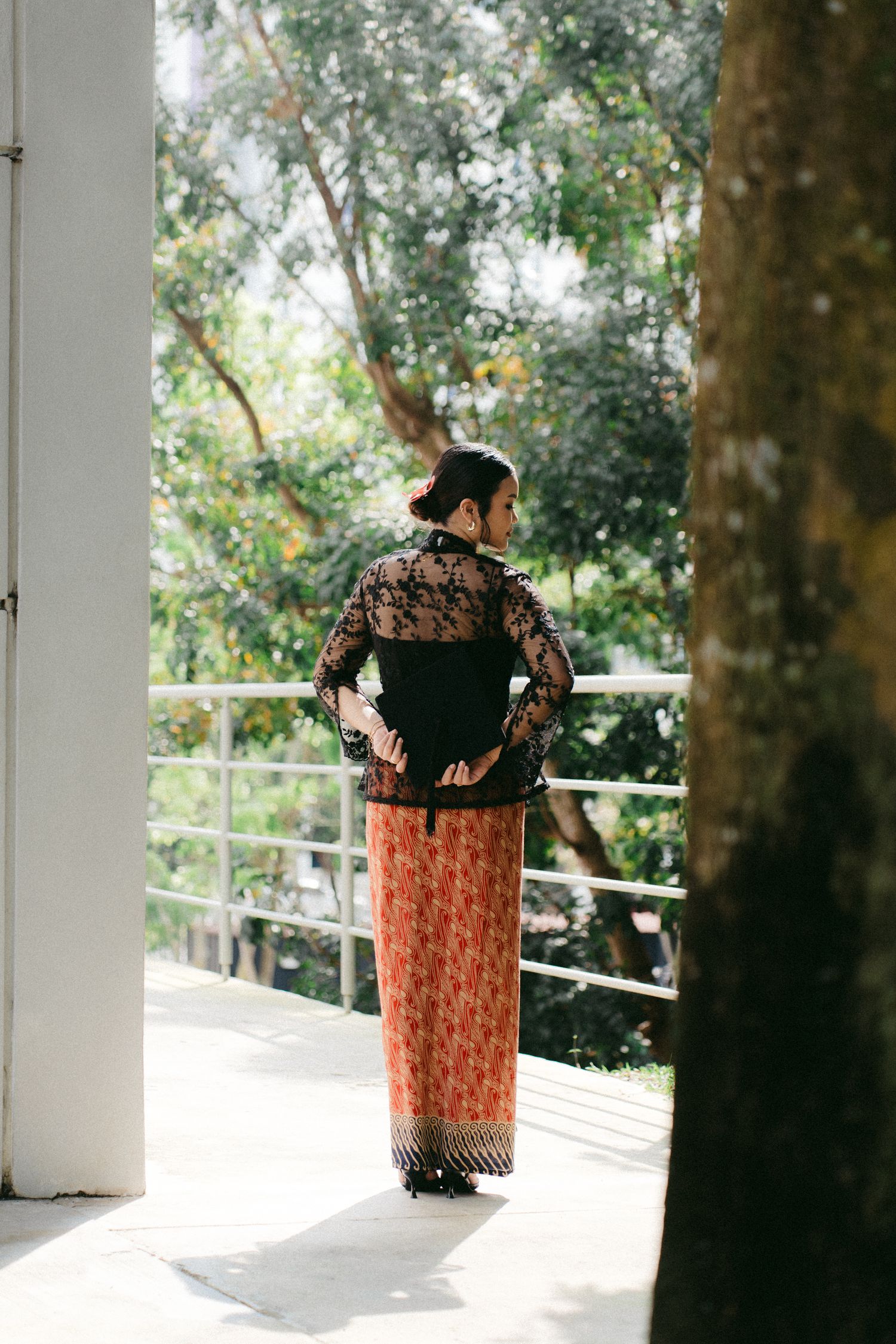 A person stands outdoors wearing a black lace top and patterned orange batik skirt against a natural background.