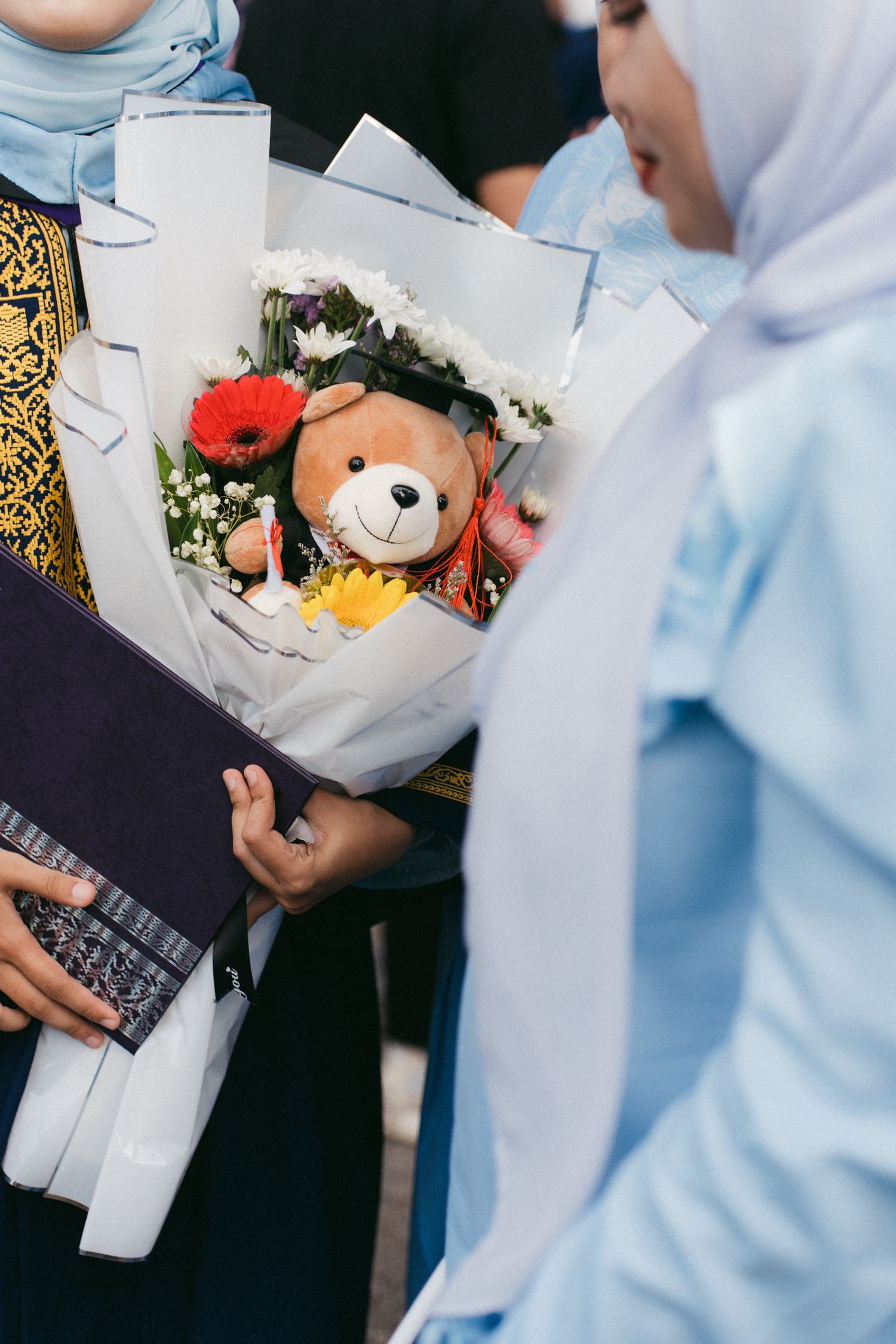 A floral bouquet with a cute teddy bear centerpiece being held, featuring red and yellow flowers wrapped in white paper.