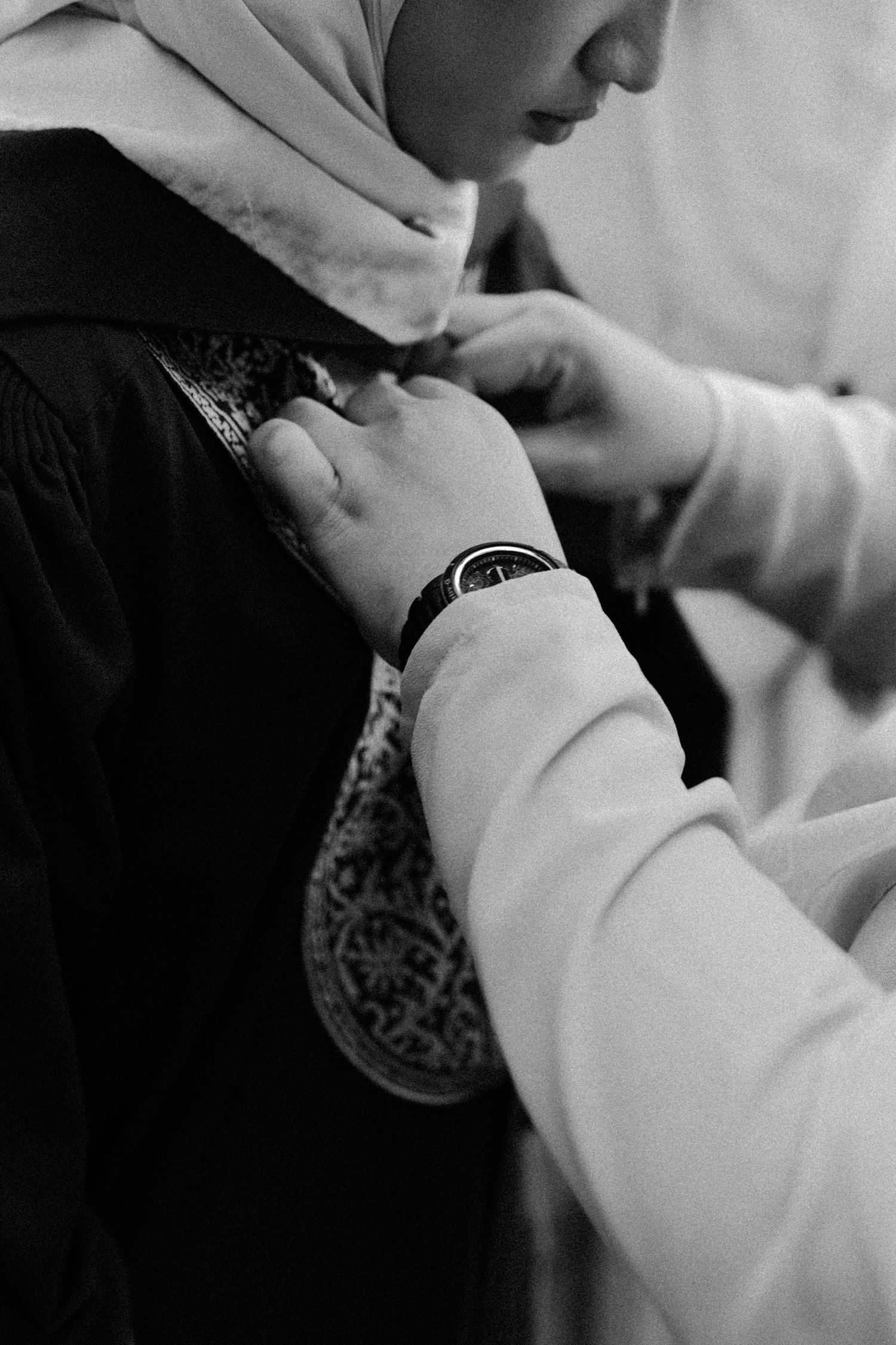 Black and white photograph of hands adjusting a patterned head covering in an intimate portrait.