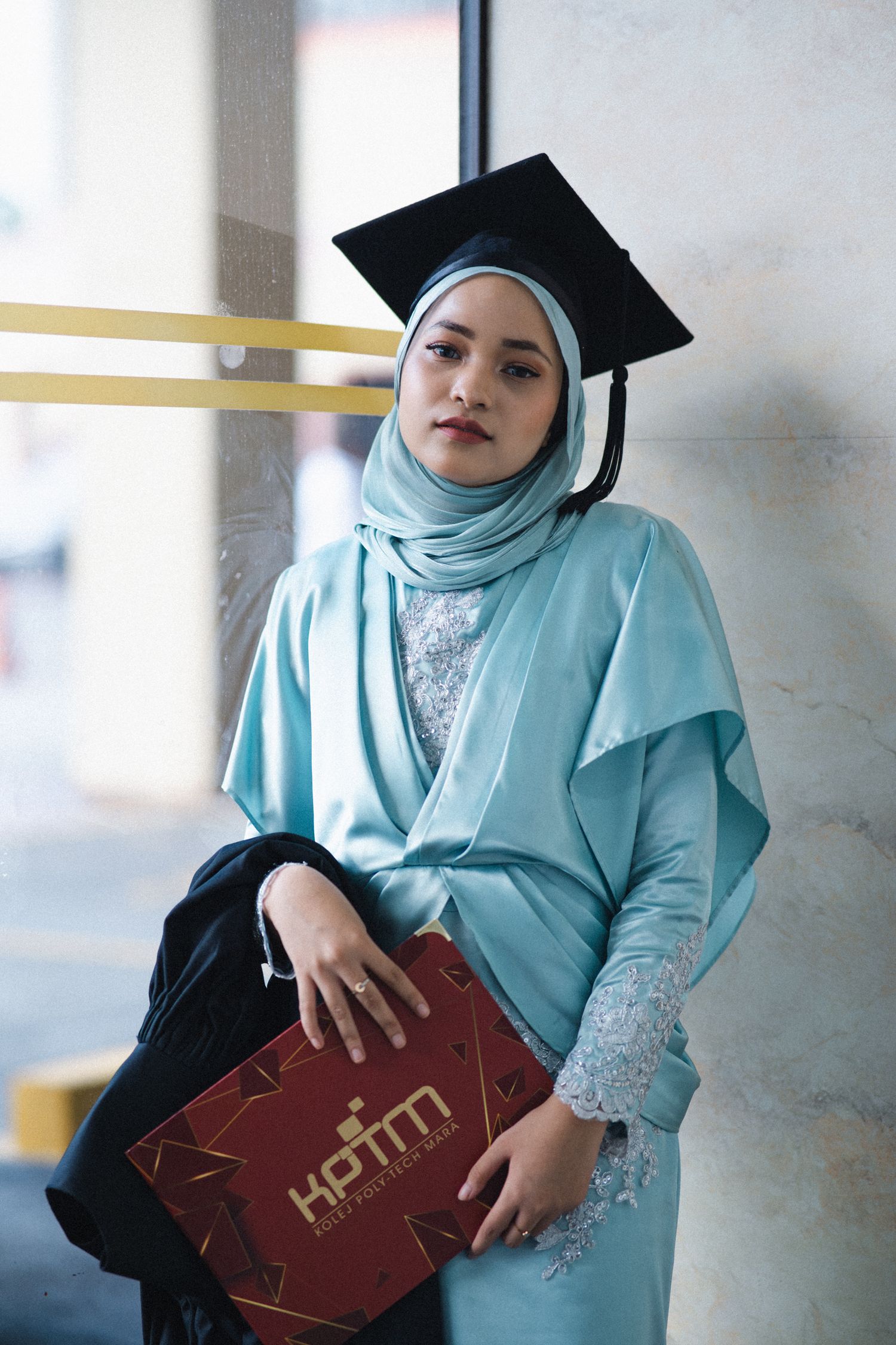 A graduate in turquoise attire poses with academic folder against a light wall.