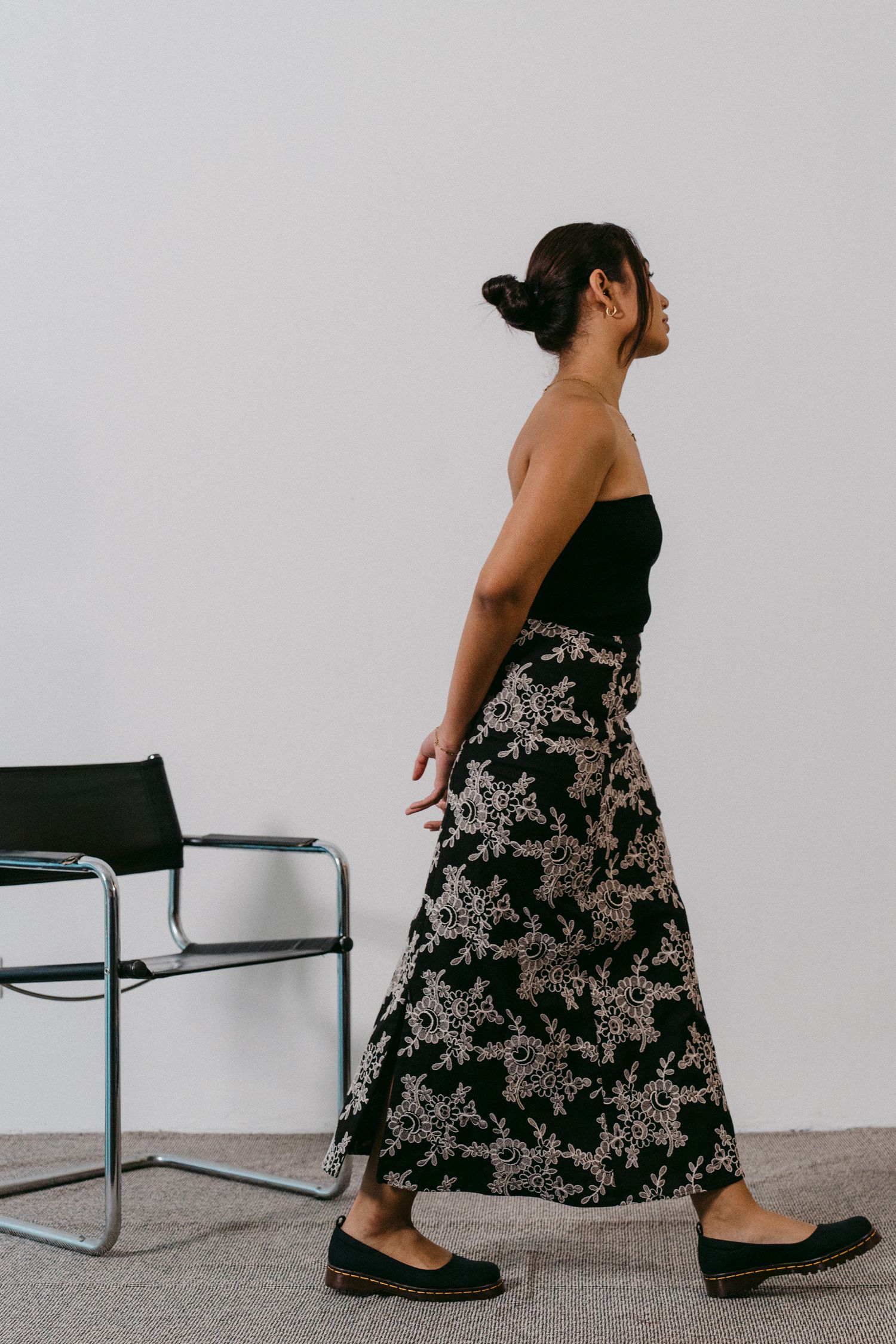 Woman wearing black top and floral maxi skirt walks past modern chair against white wall.
