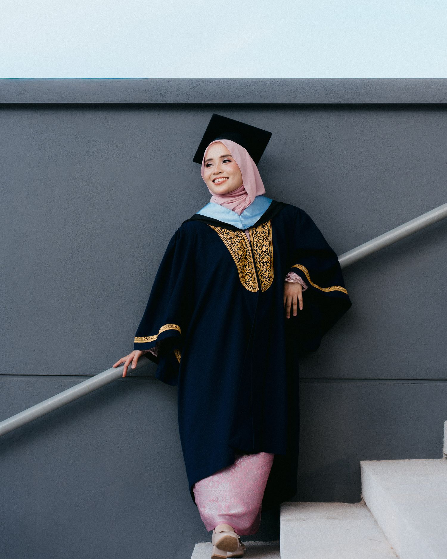 Graduate in navy academic regalia with gold embellishments poses on stairs against grey wall.