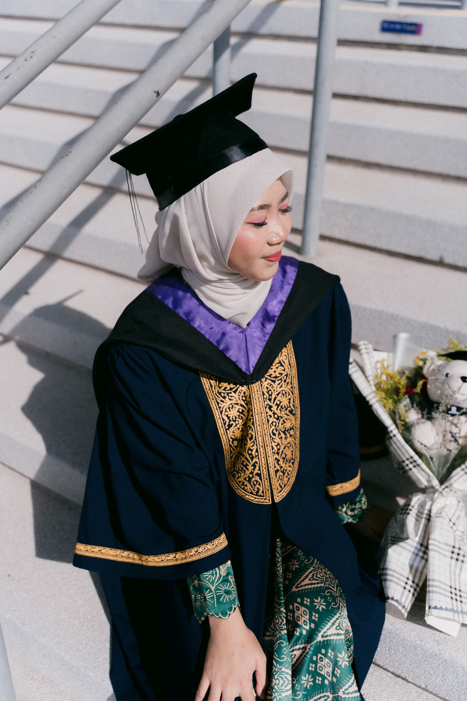 A person wearing a black graduation gown with gold embellishments and hijab poses on concrete steps.