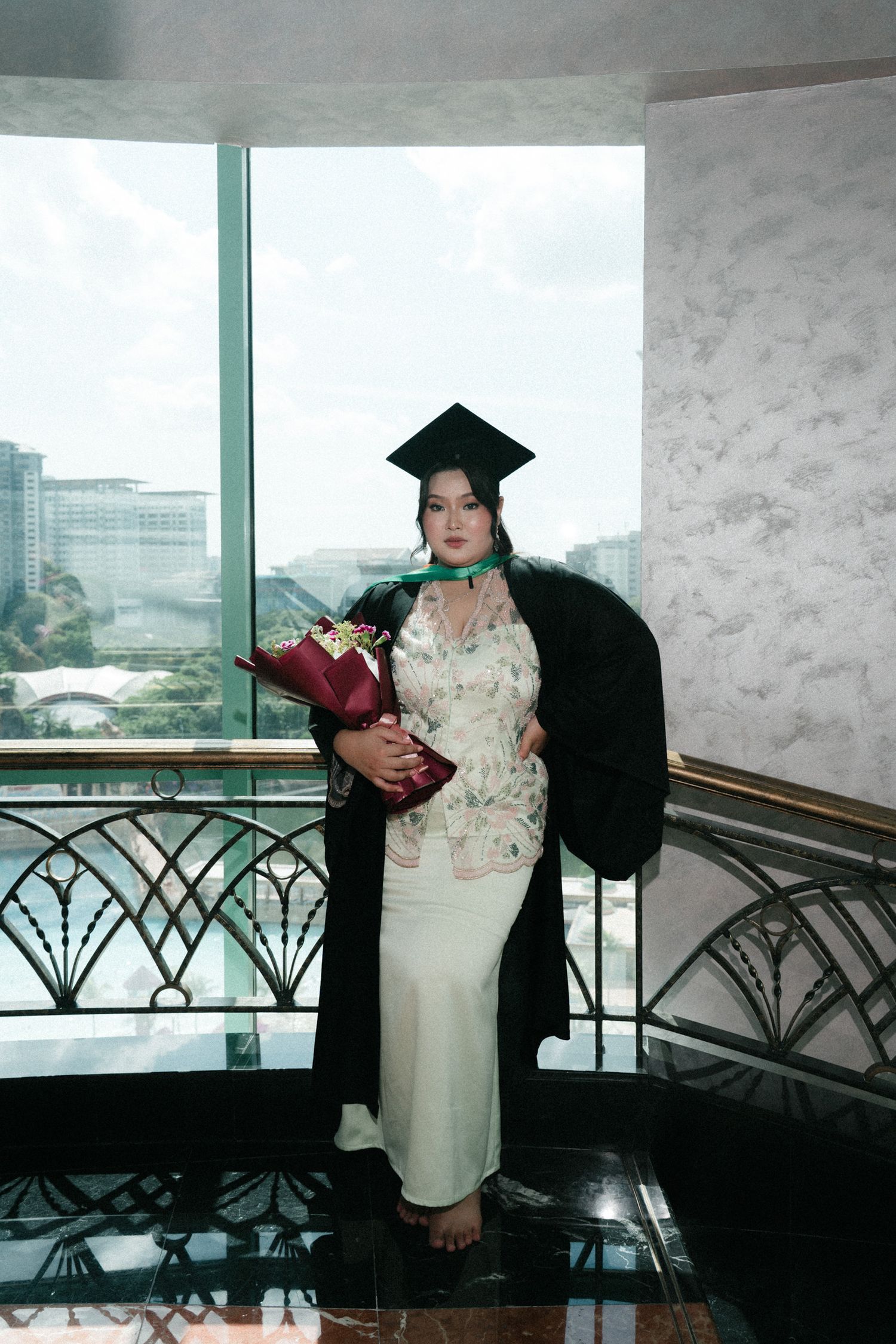 A graduate in black cap and gown poses near a decorative railing in a modern building with city views.