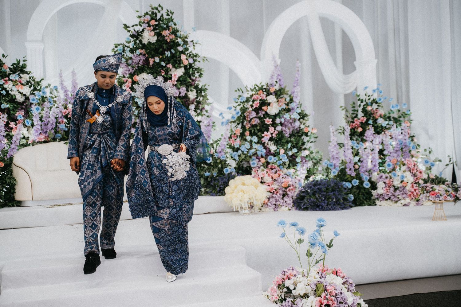 A couple in matching navy blue traditional attire walk together at a wedding venue decorated with floral arrangements.