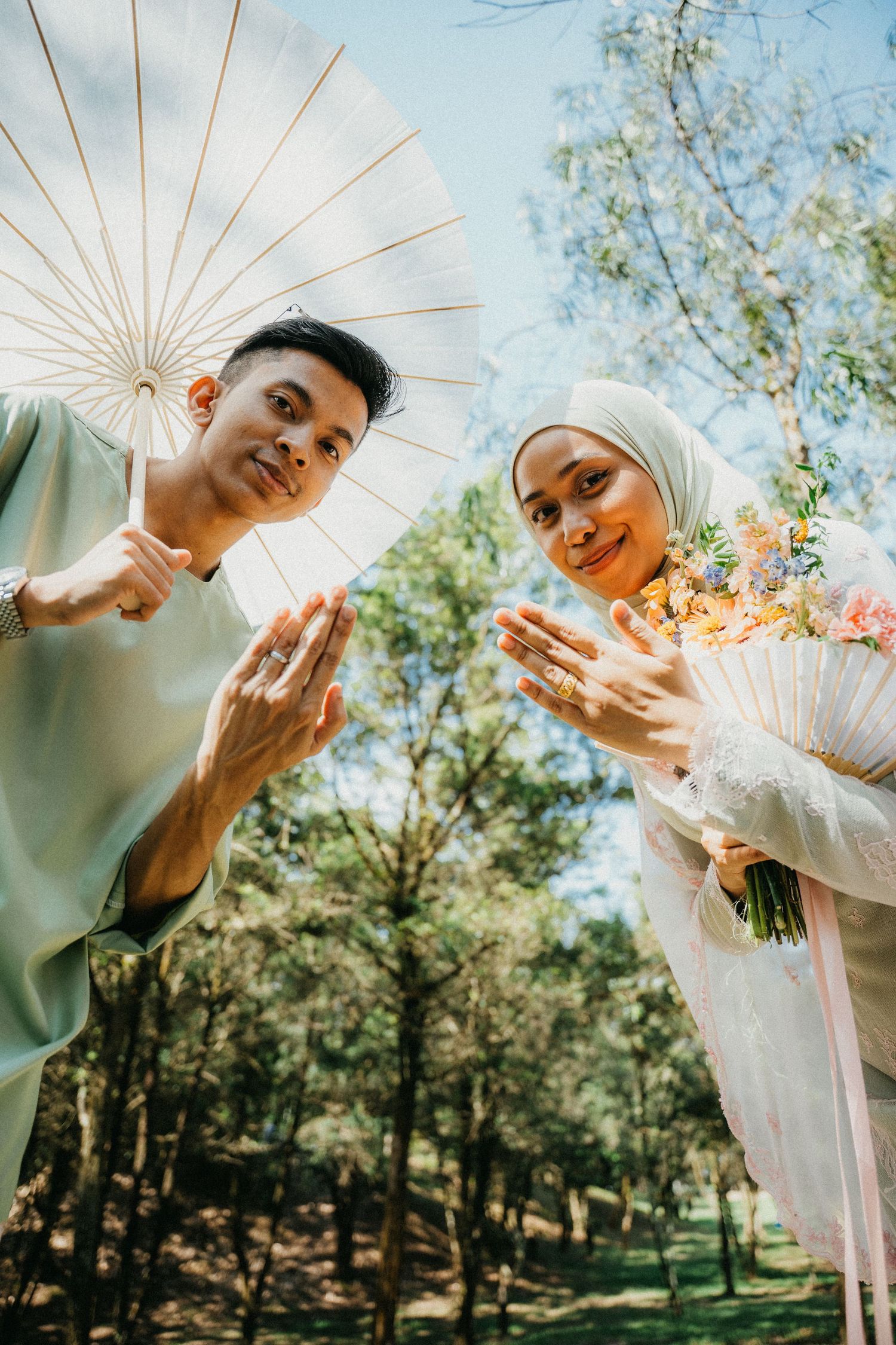 Low angle view of couple in light clothing outdoors with parasol and bouquet against green forest background.