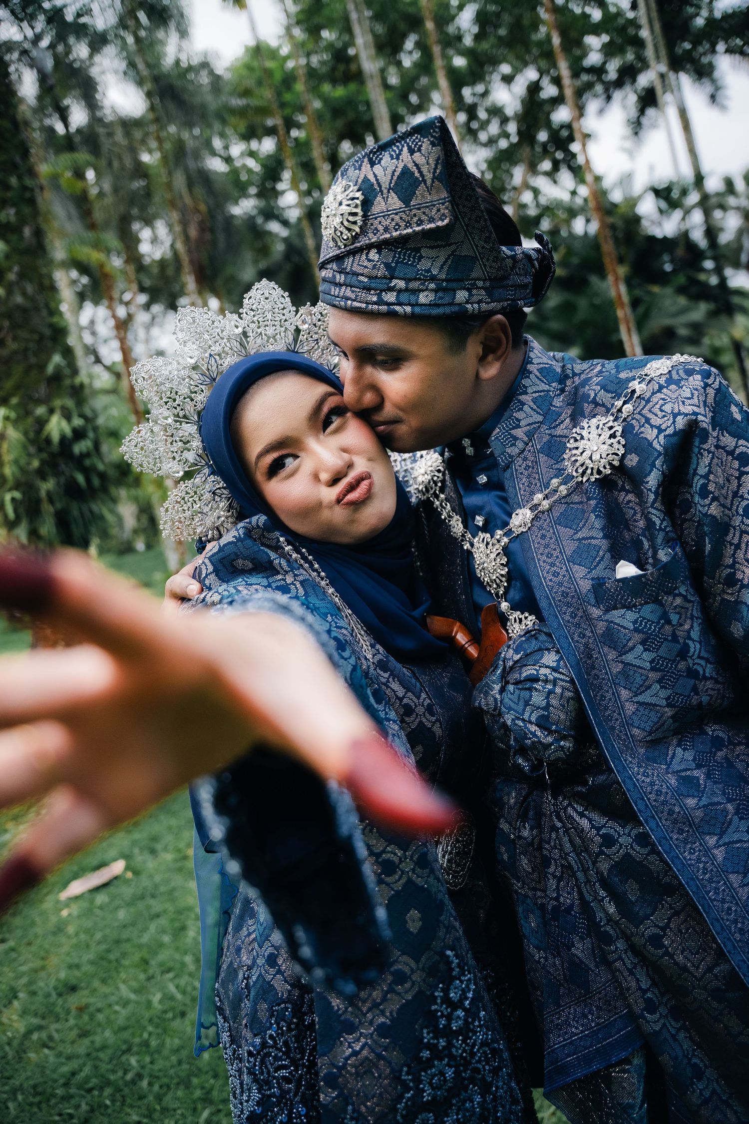 Traditional Malay wedding couple in matching ornate blue and silver traditional attire embracing in garden setting.