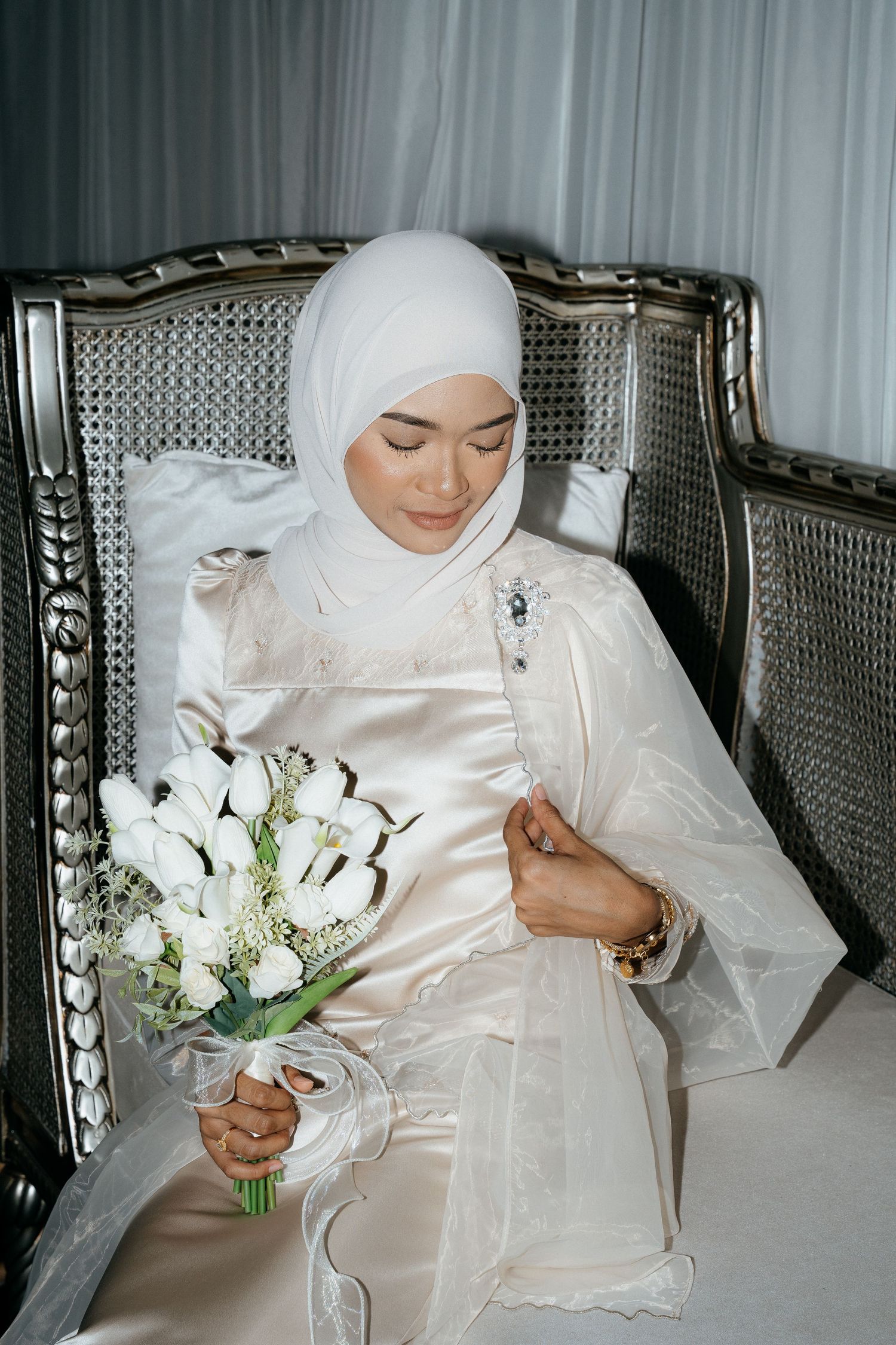 A bride in white hijab and silk dress holding a delicate bouquet of white flowers while sitting in an elegant chair.