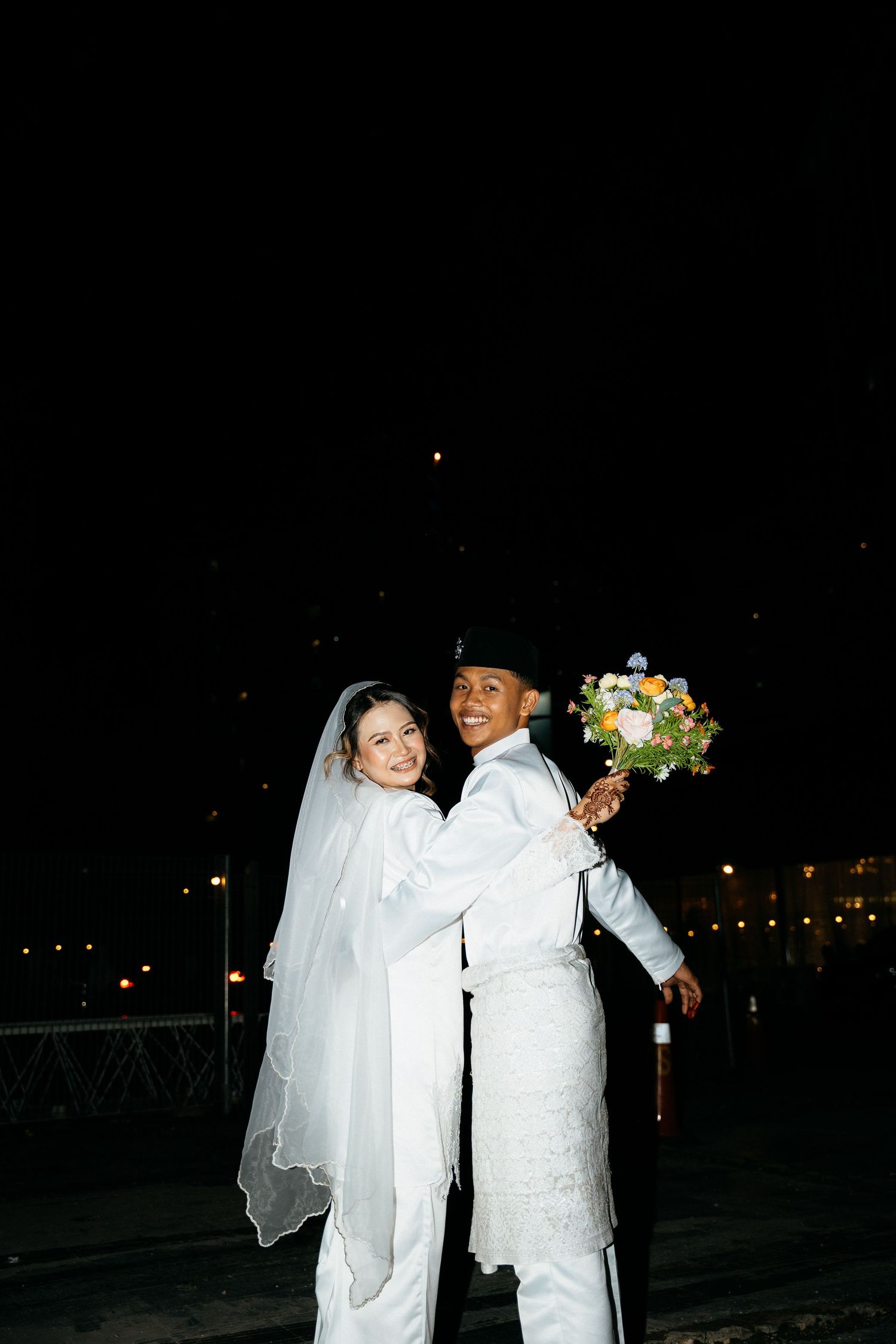 A newlywed couple in white attire shares a joyful moment at night while holding a colorful floral bouquet.