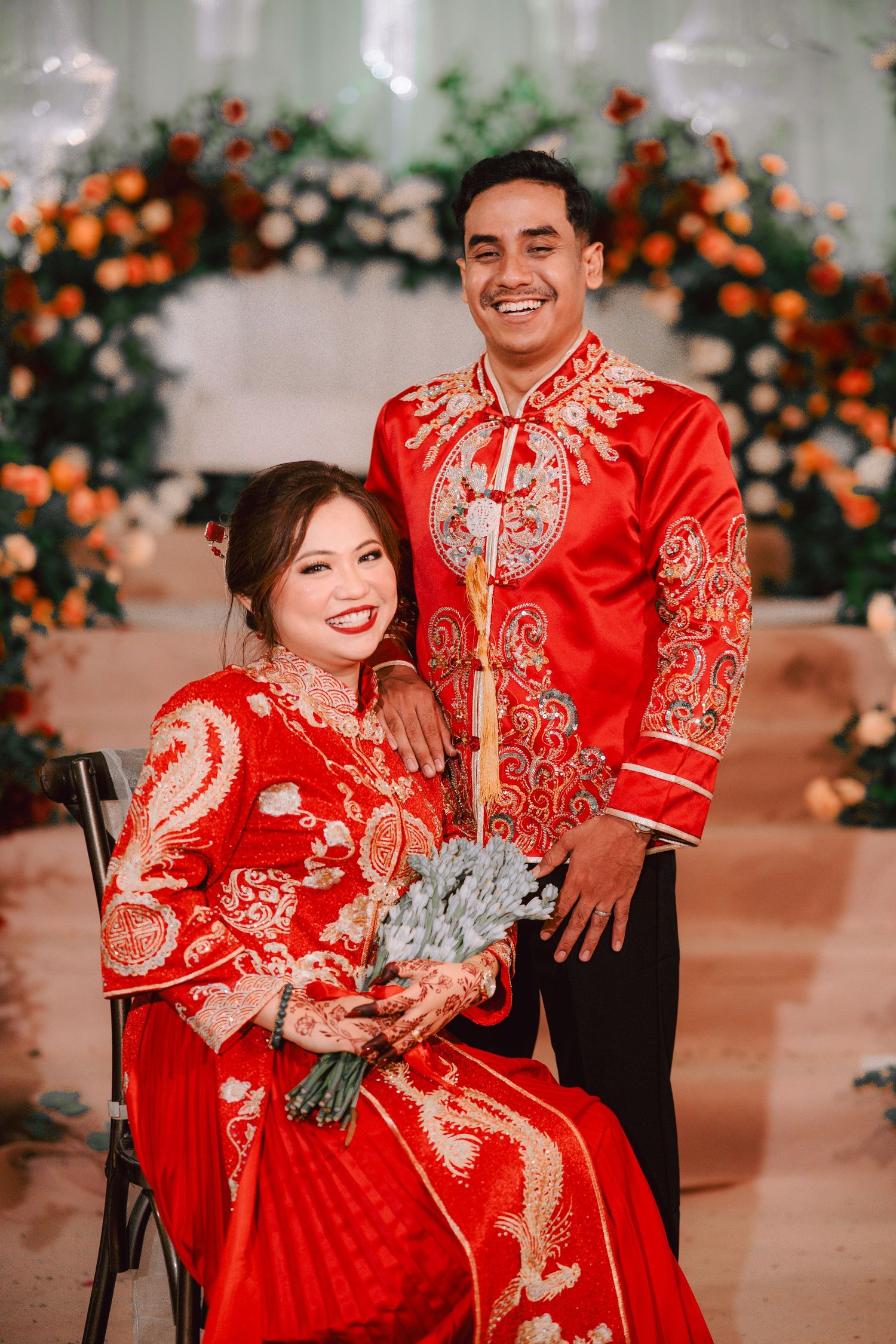 A couple in traditional Chinese wedding attire with red embroidered outfits posing together during their celebration ceremony.