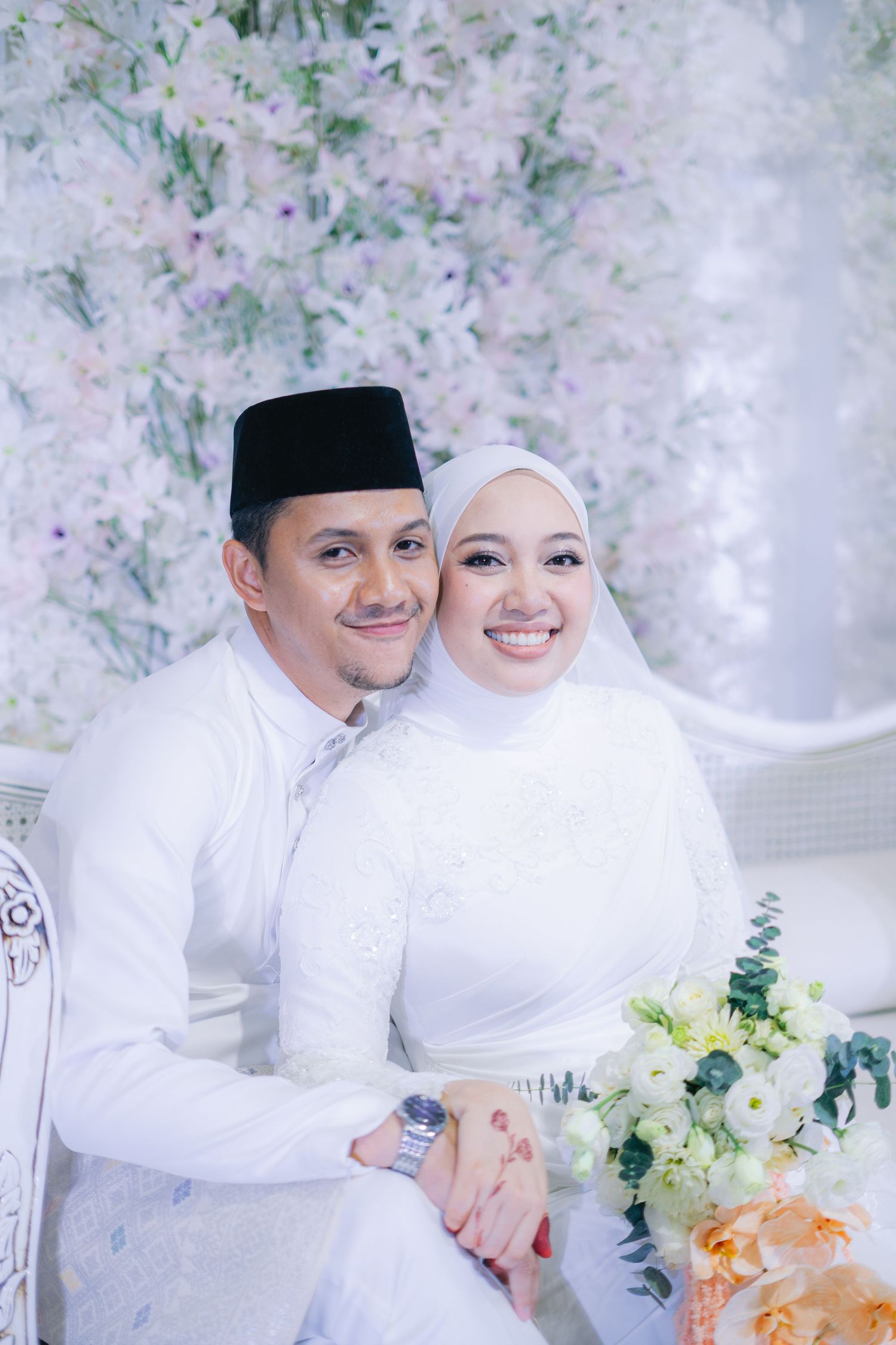A happy couple in traditional white Malay wedding attire poses together against a floral backdrop during their celebration.