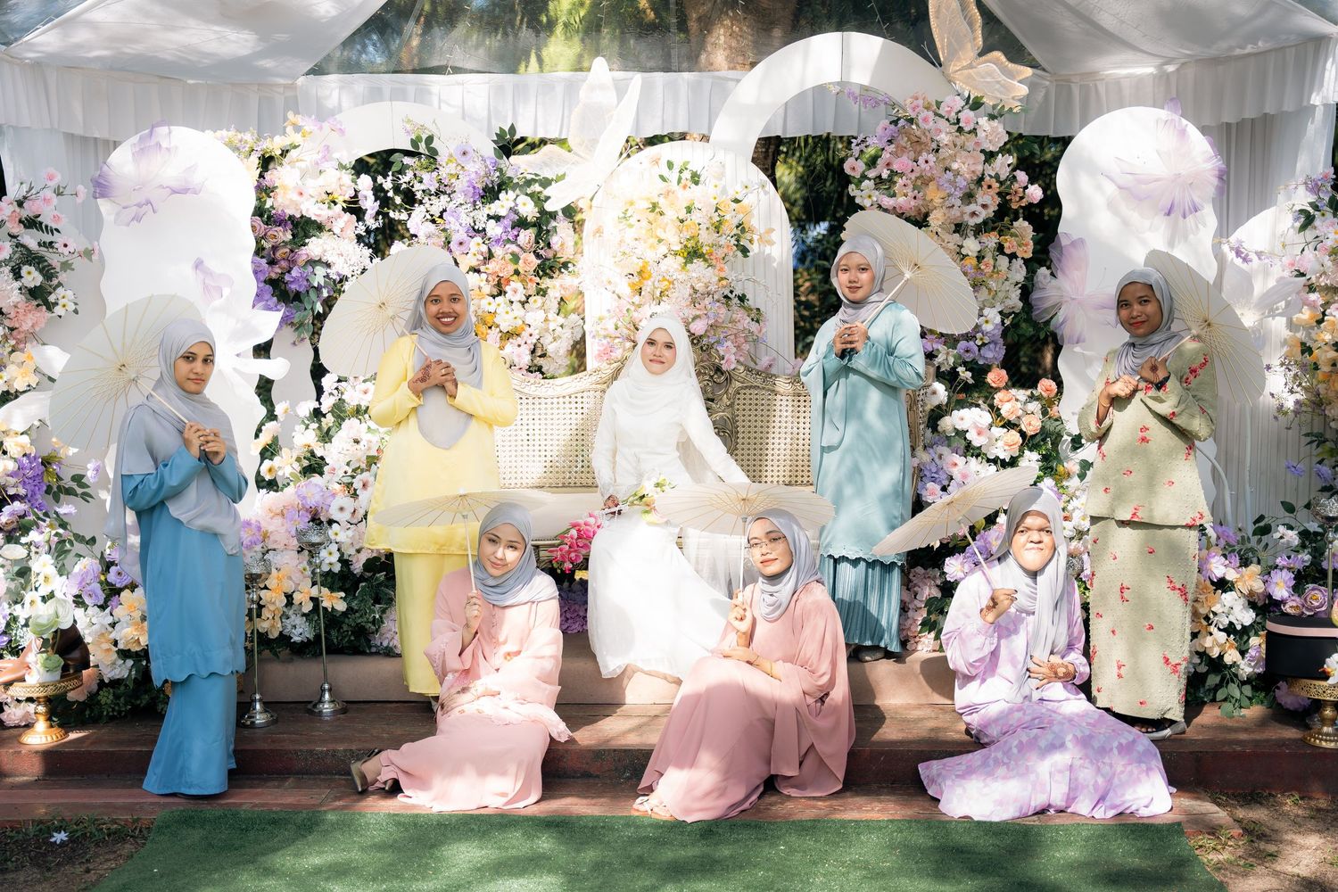A group in pastel hijabs and traditional outfits pose together in front of a floral arch at a wedding celebration.