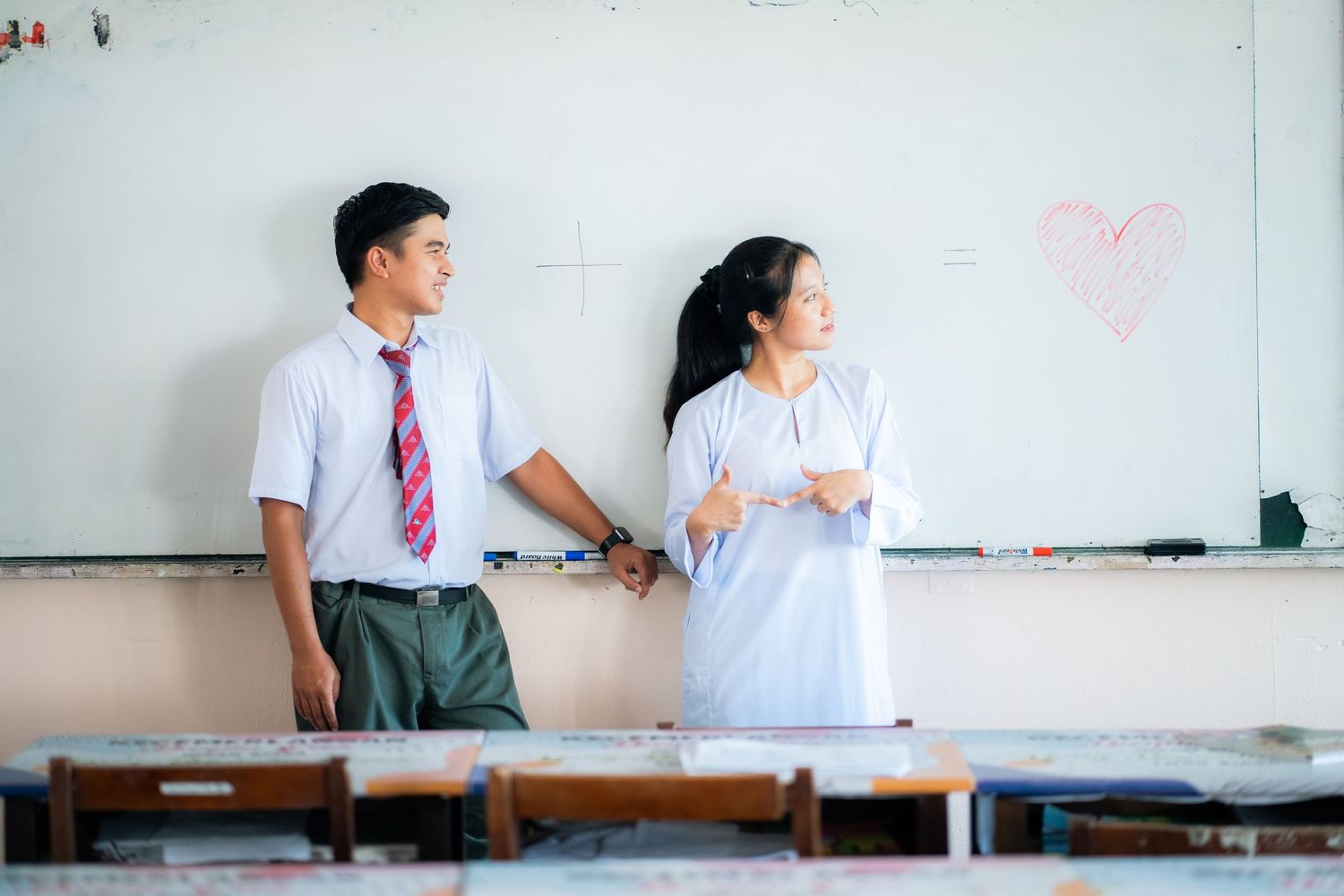 Students in school uniforms share a lighthearted moment in front of classroom wall with pink heart drawing.