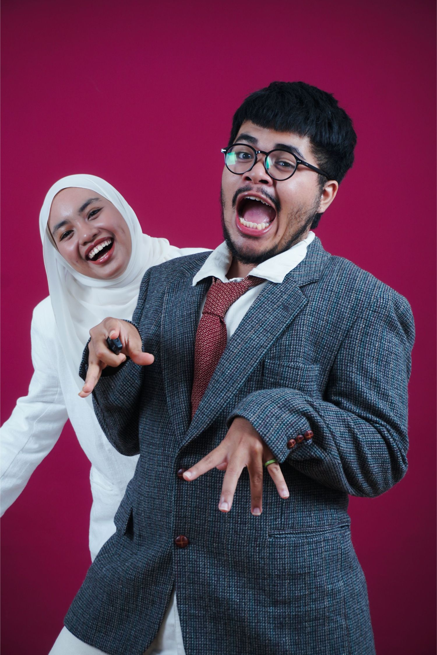 Two young Southeast Asian people laughing and posing expressively against a vibrant pink background in formal attire.