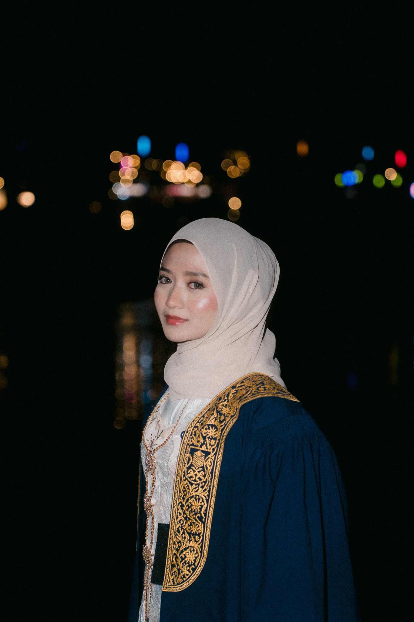Young Muslim woman in hijab and ornate blue robe poses outdoors at night with bokeh city lights behind her.