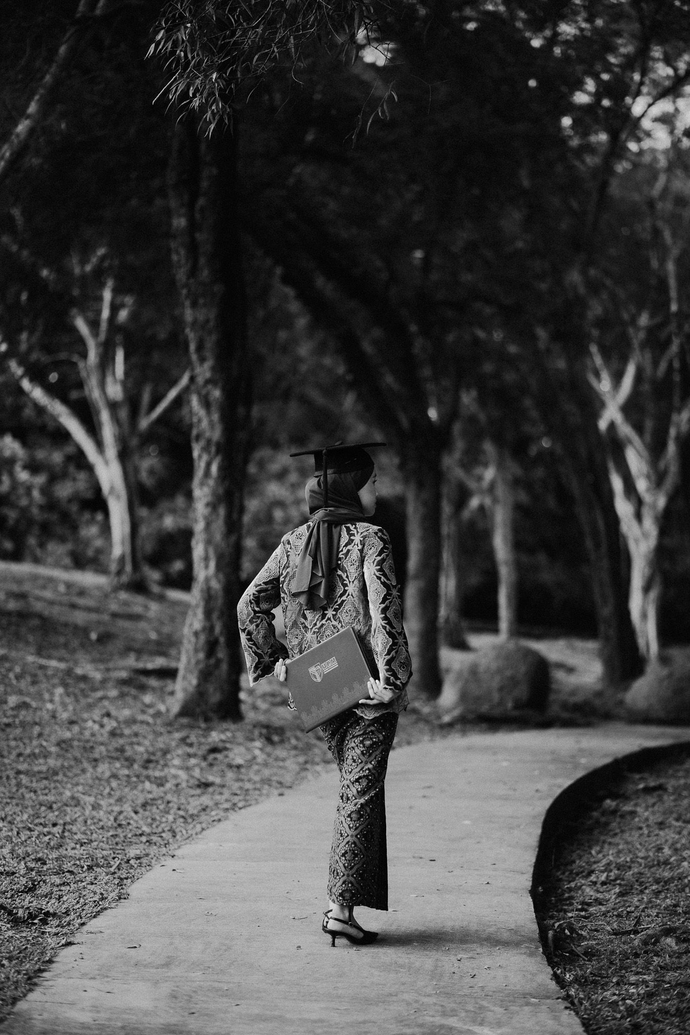 Black and white photo of a graduate in cap and gown holding a diploma, walking along a tree-lined path.