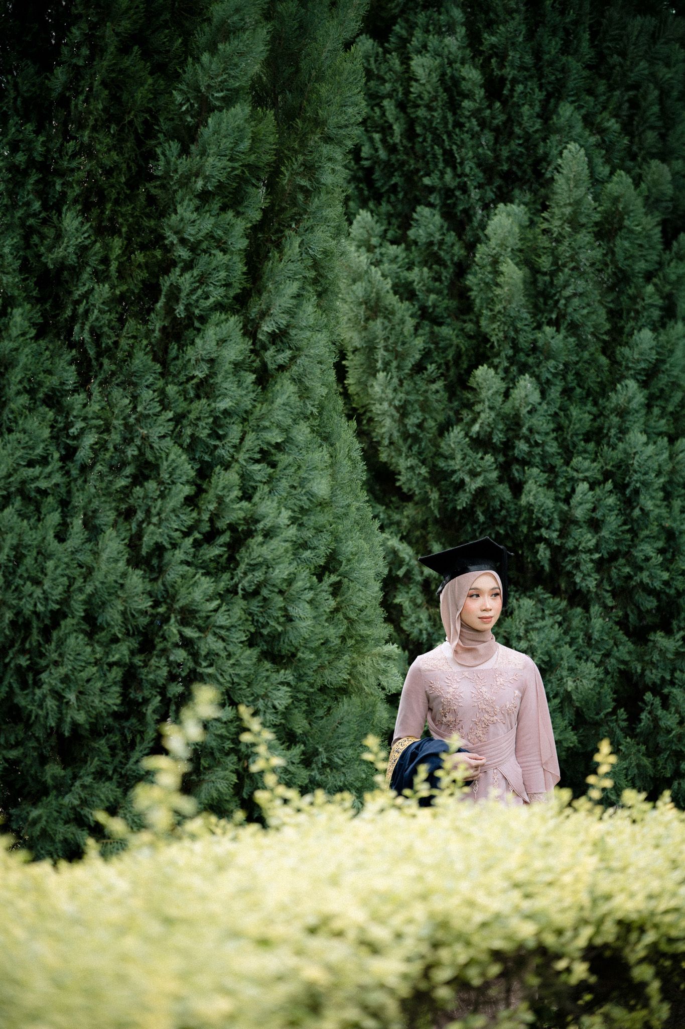 A graduate in hijab and cap poses among lush green cypress trees and yellow flowers outdoors.
