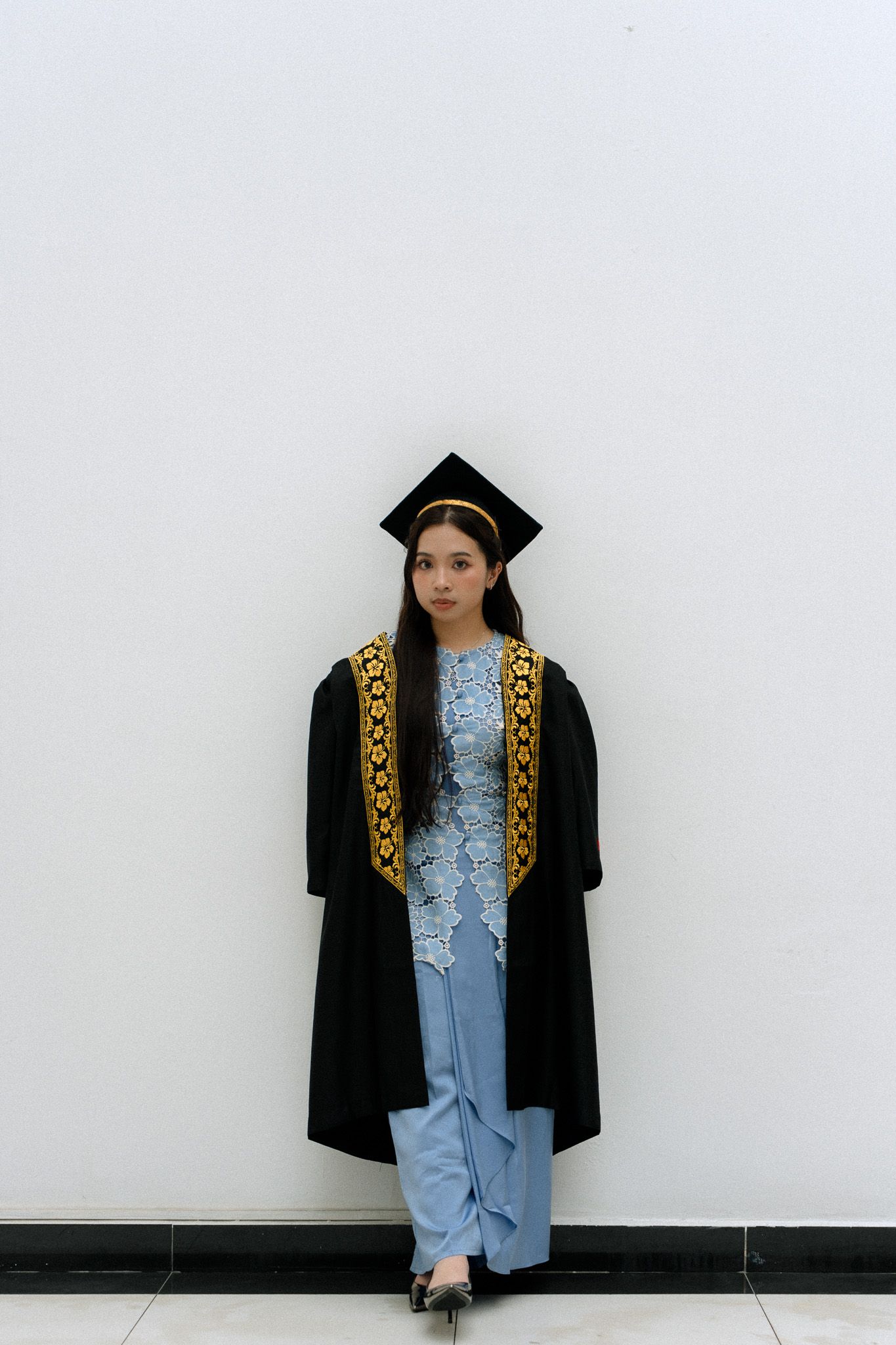 Young woman in black graduation gown and cap with gold trim poses confidently against a white wall.