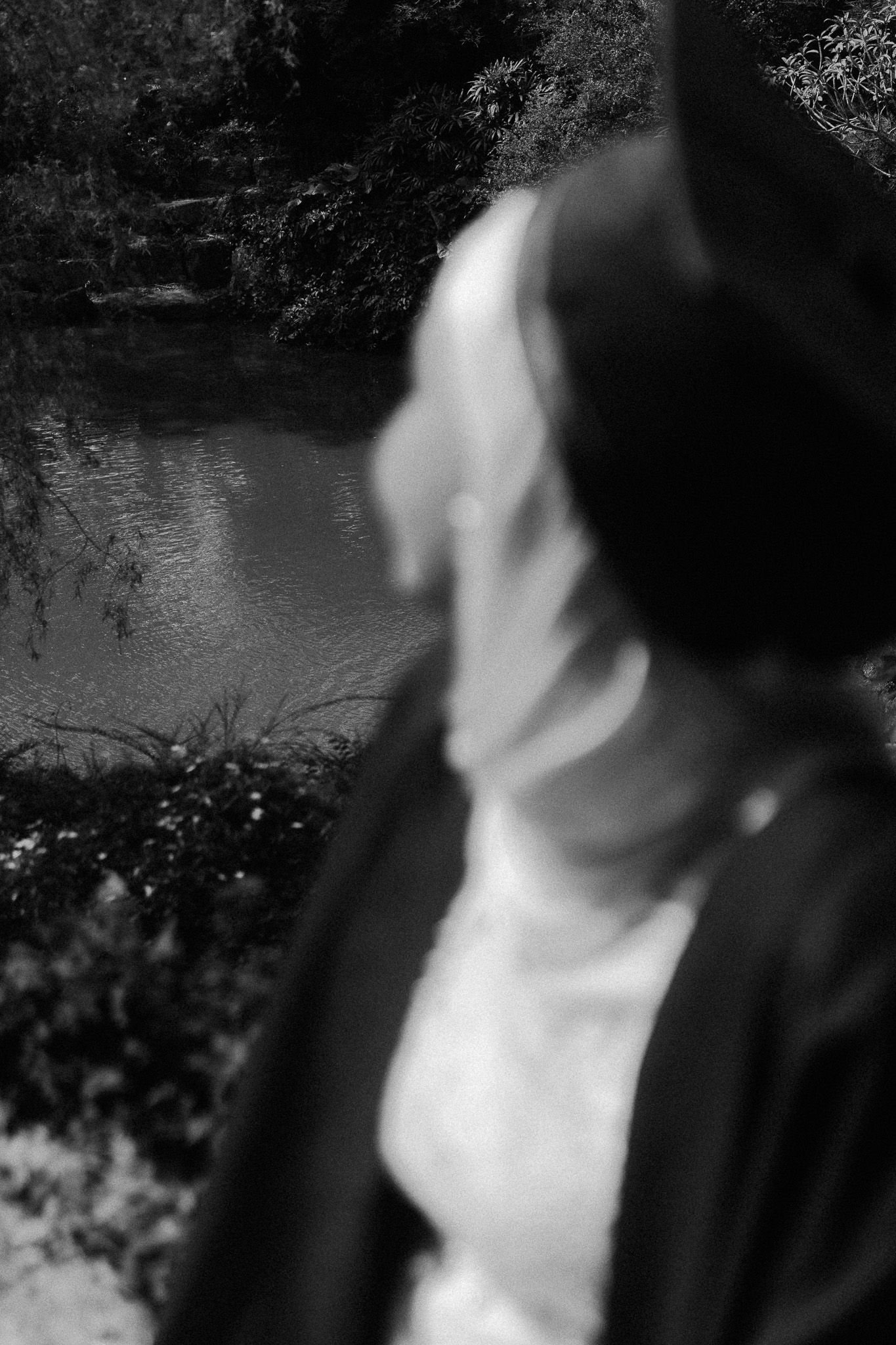Close-up black and white portrait of a person wearing a hat and white scarf near a reflective pond in a garden.