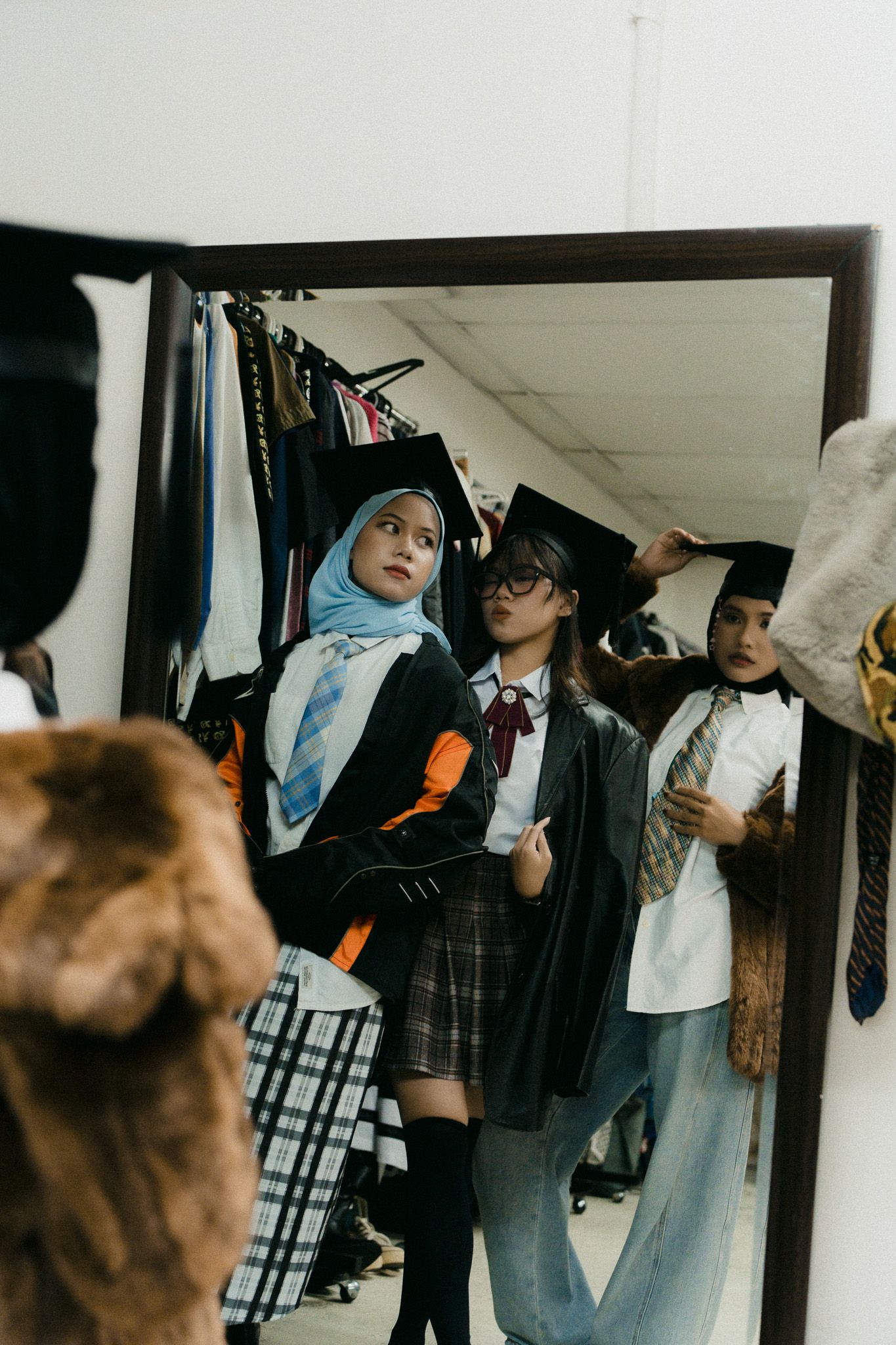 Two women in academic caps try on outfits in a clothing store mirror reflection.