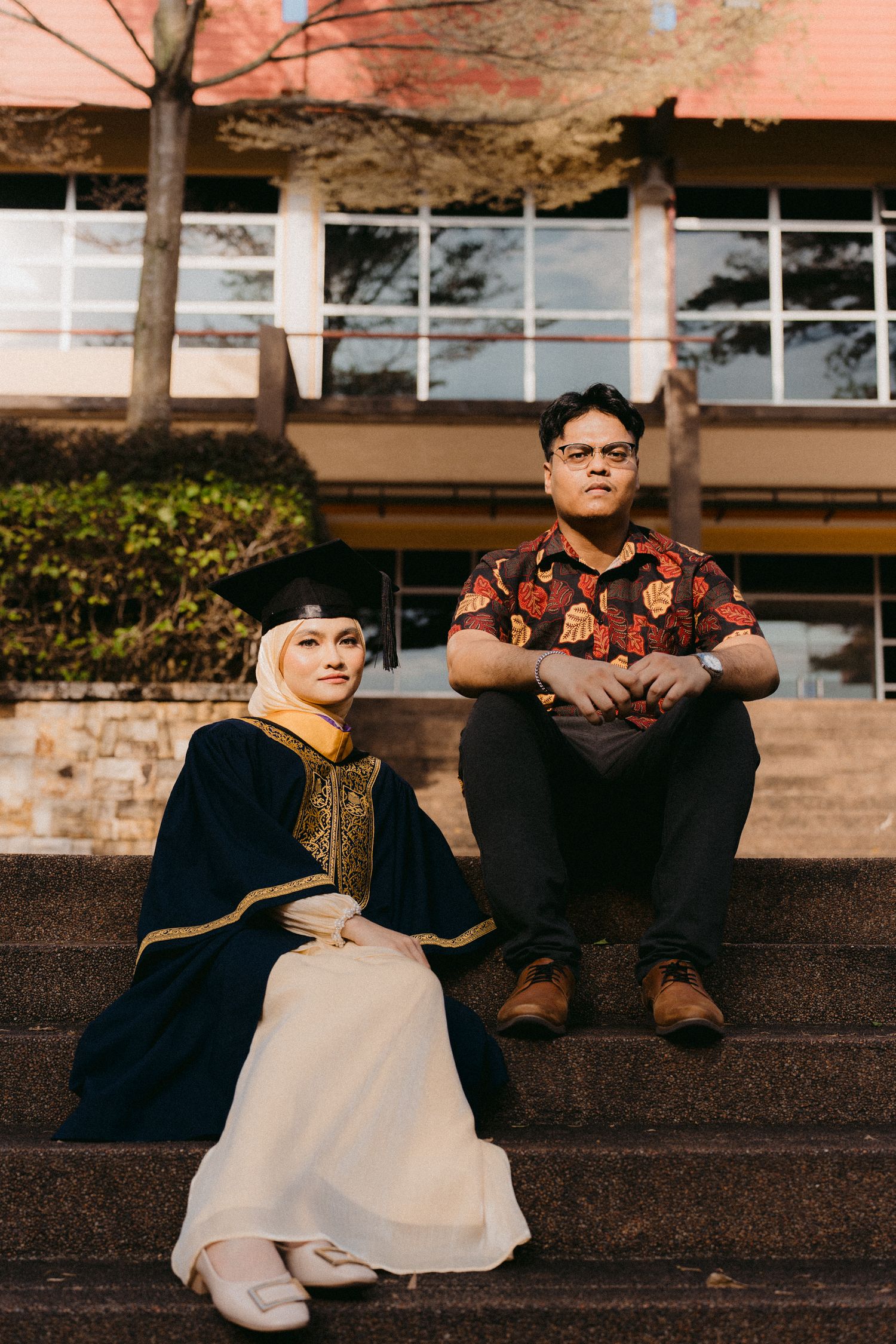 Two people sitting on concrete steps outside a building with windows on a warm-toned autumn day.