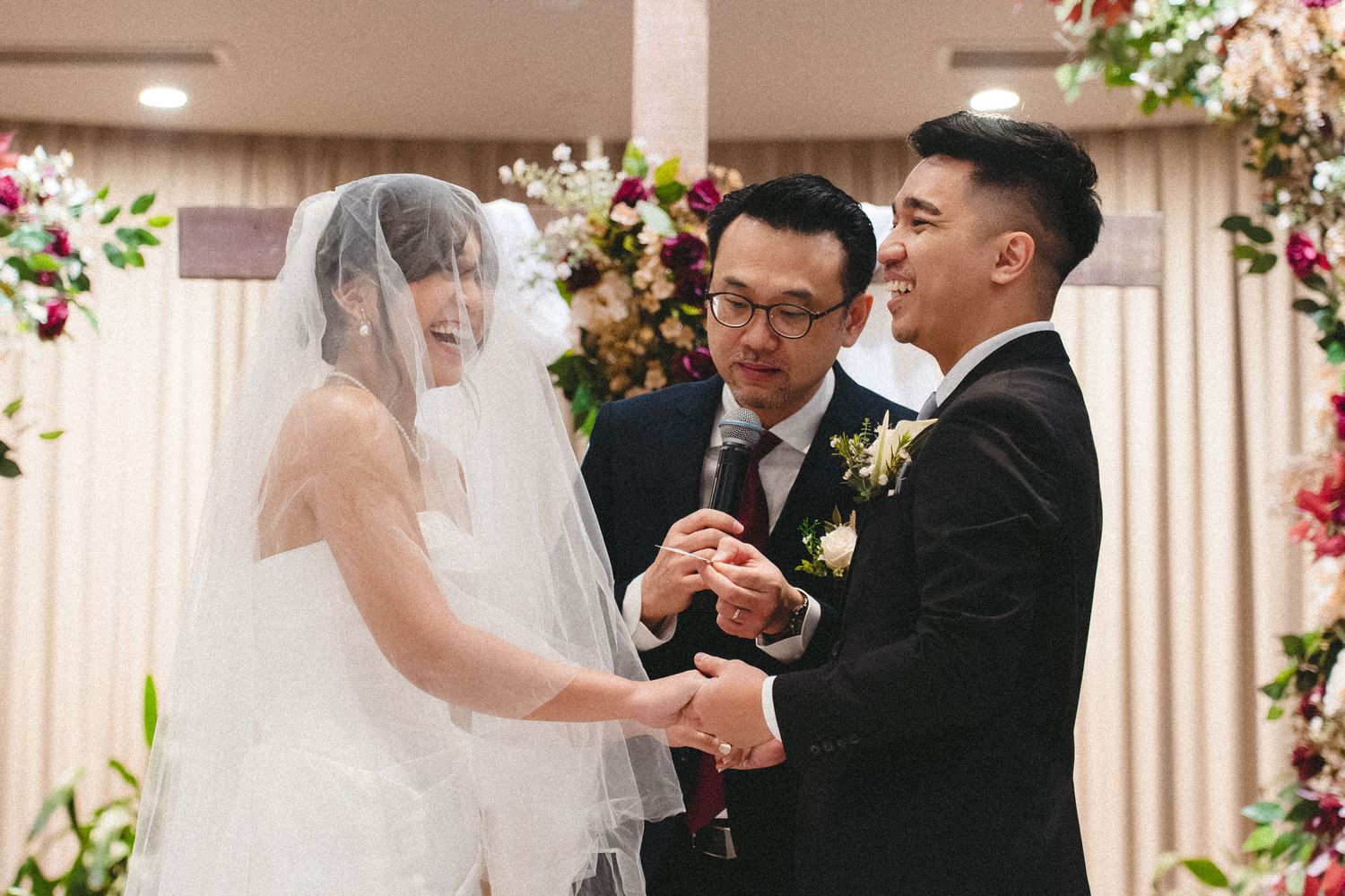 Wedding ceremony taking place indoors with floral decor and a veiled bride exchanging rings during the vows.