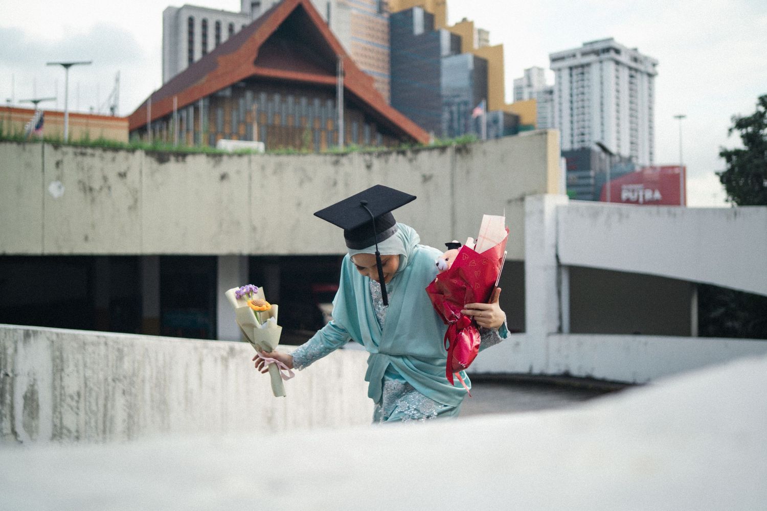 Graduate in teal dress and cap sits casually on concrete wall with city buildings in background.