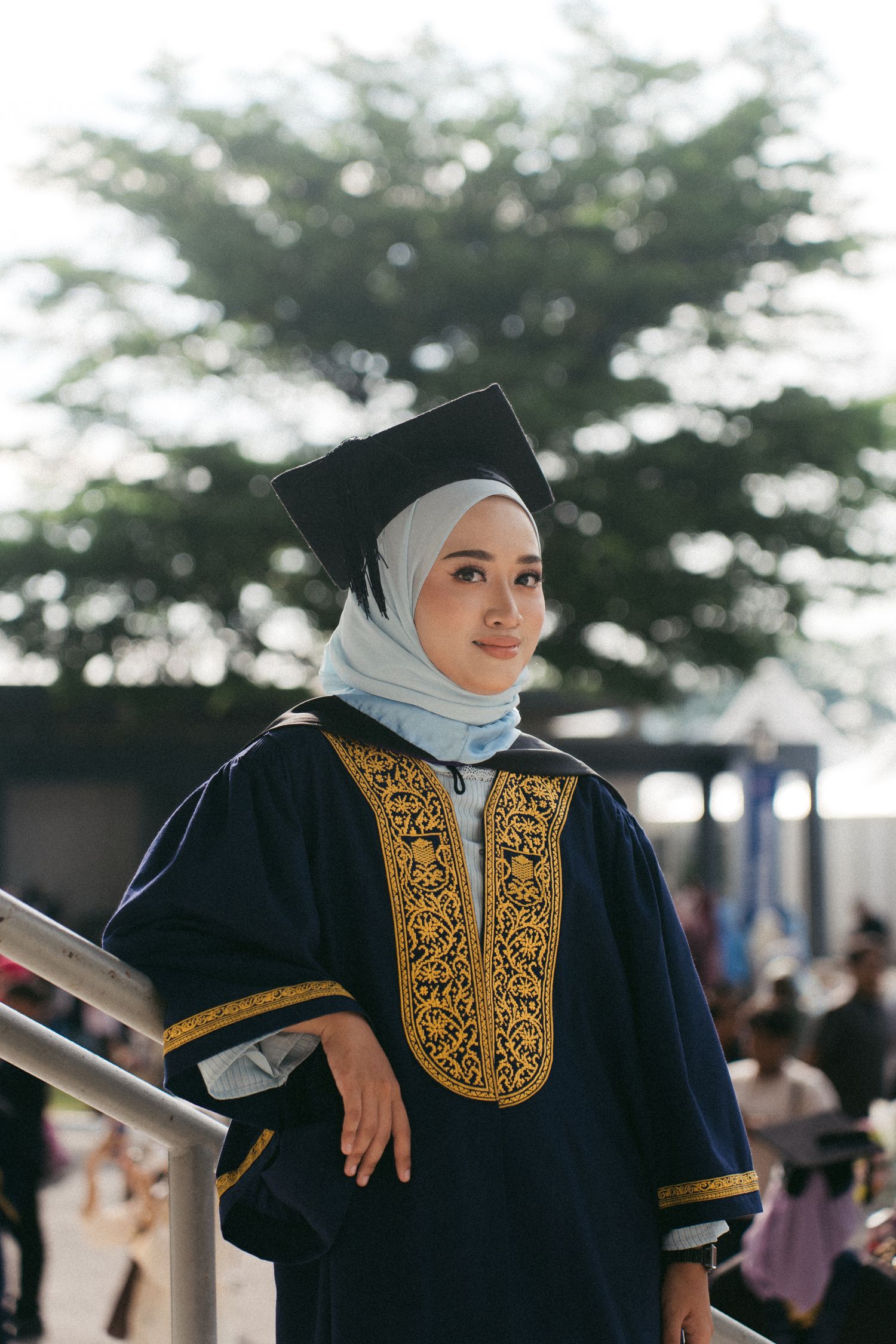 A graduate in traditional academic regalia with gold embroidery poses on a staircase during commencement day.