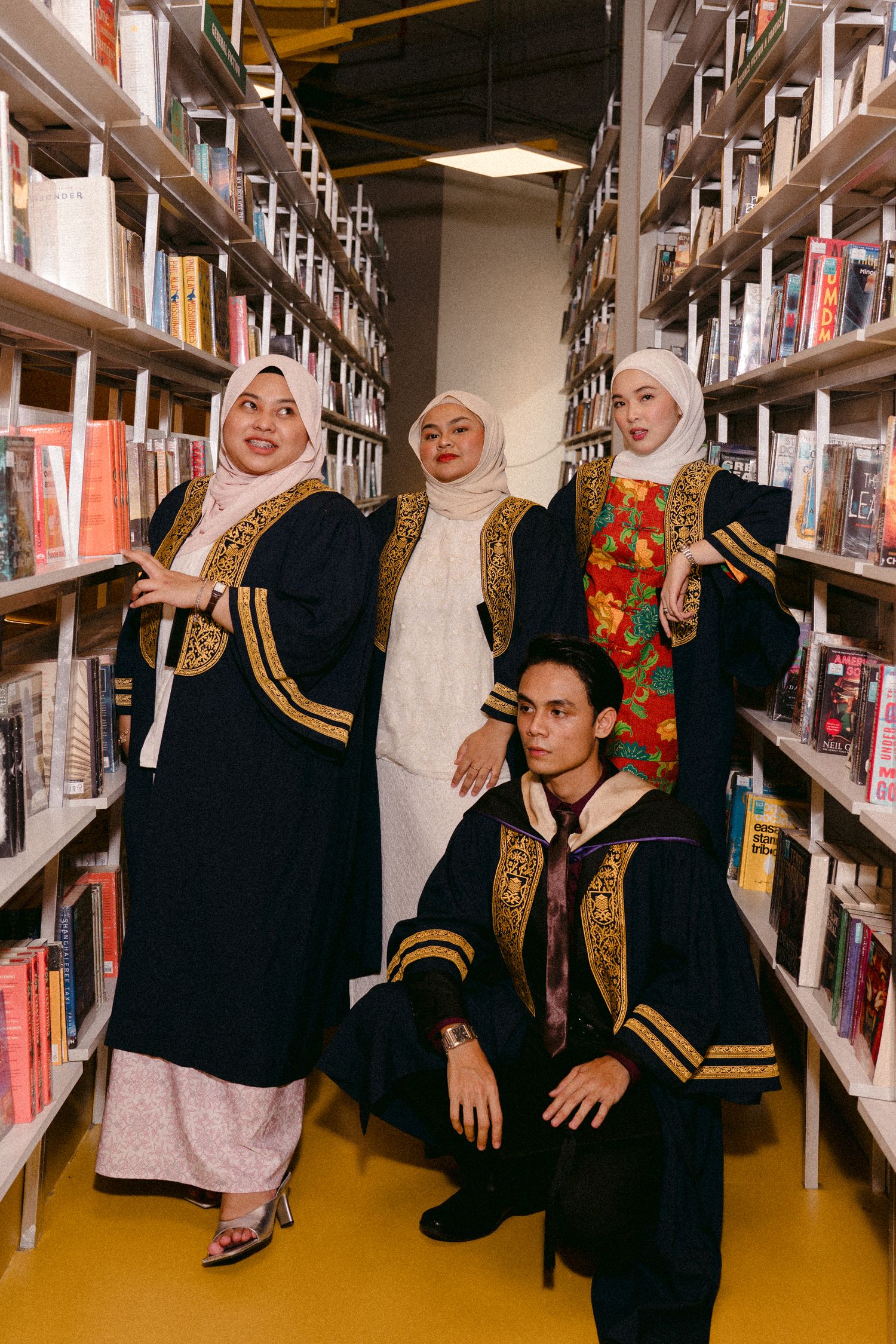 Graduates in traditional robes posing between library bookshelves for commemorative photo.
