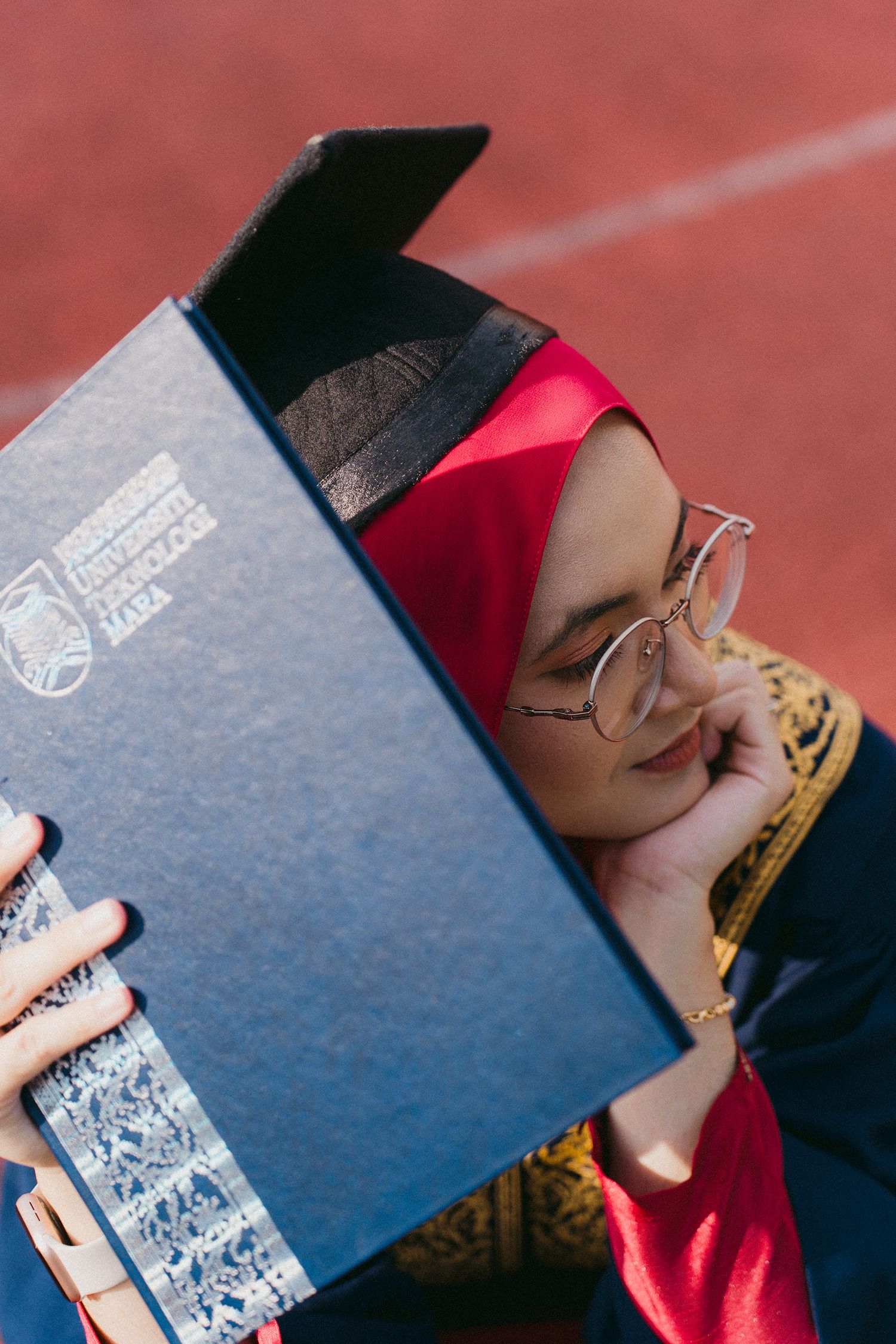 Creative shot of graduate peeking behind diploma while wearing glasses and red hijab.