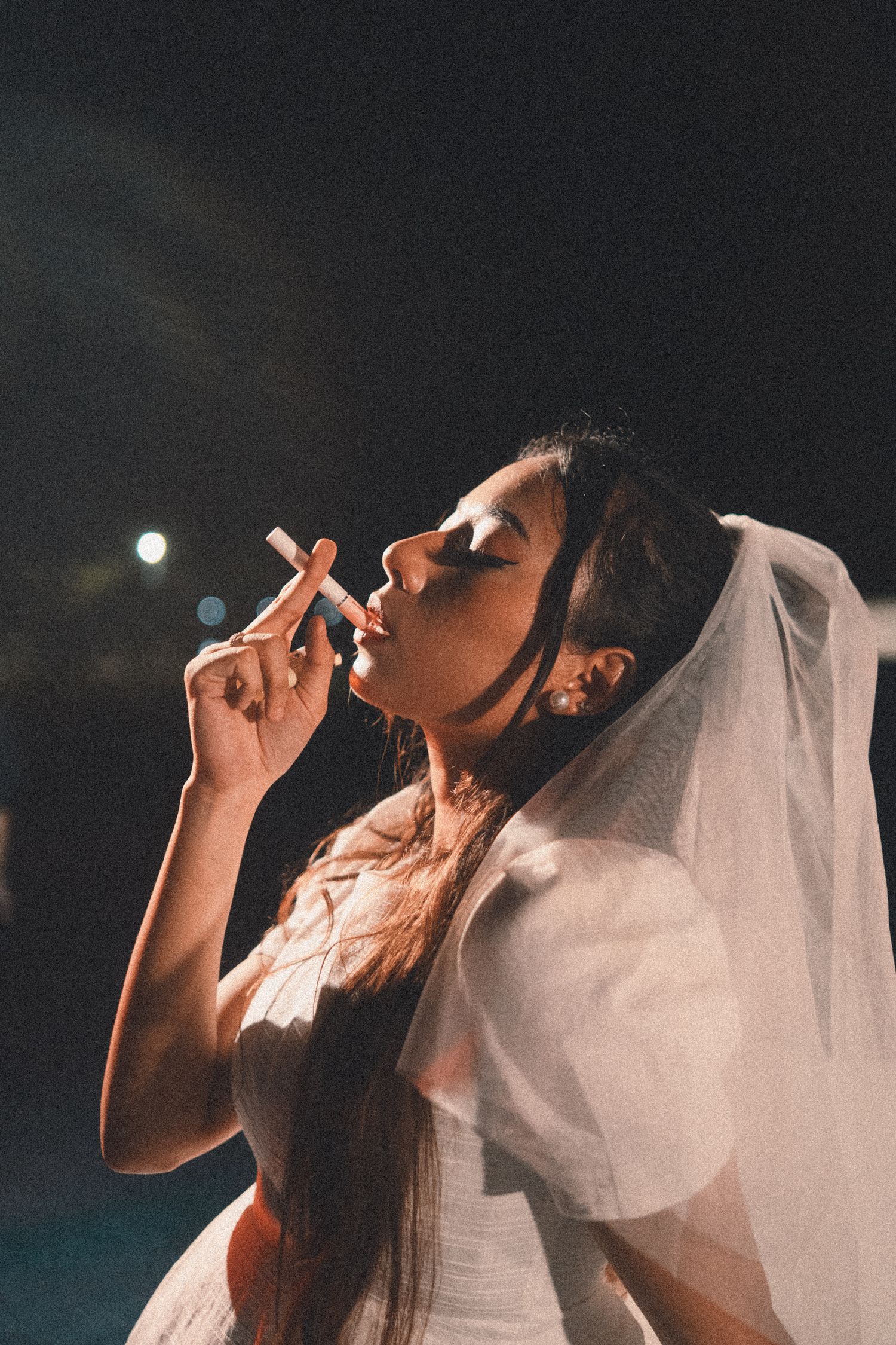 Artistic photo of person in white dress with veil at night smoking a cigarette.