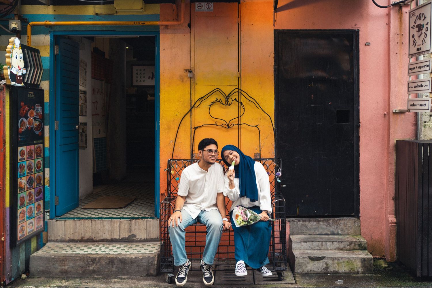 Two friends sit together on a bench against a wall with yellow heart graffiti art.