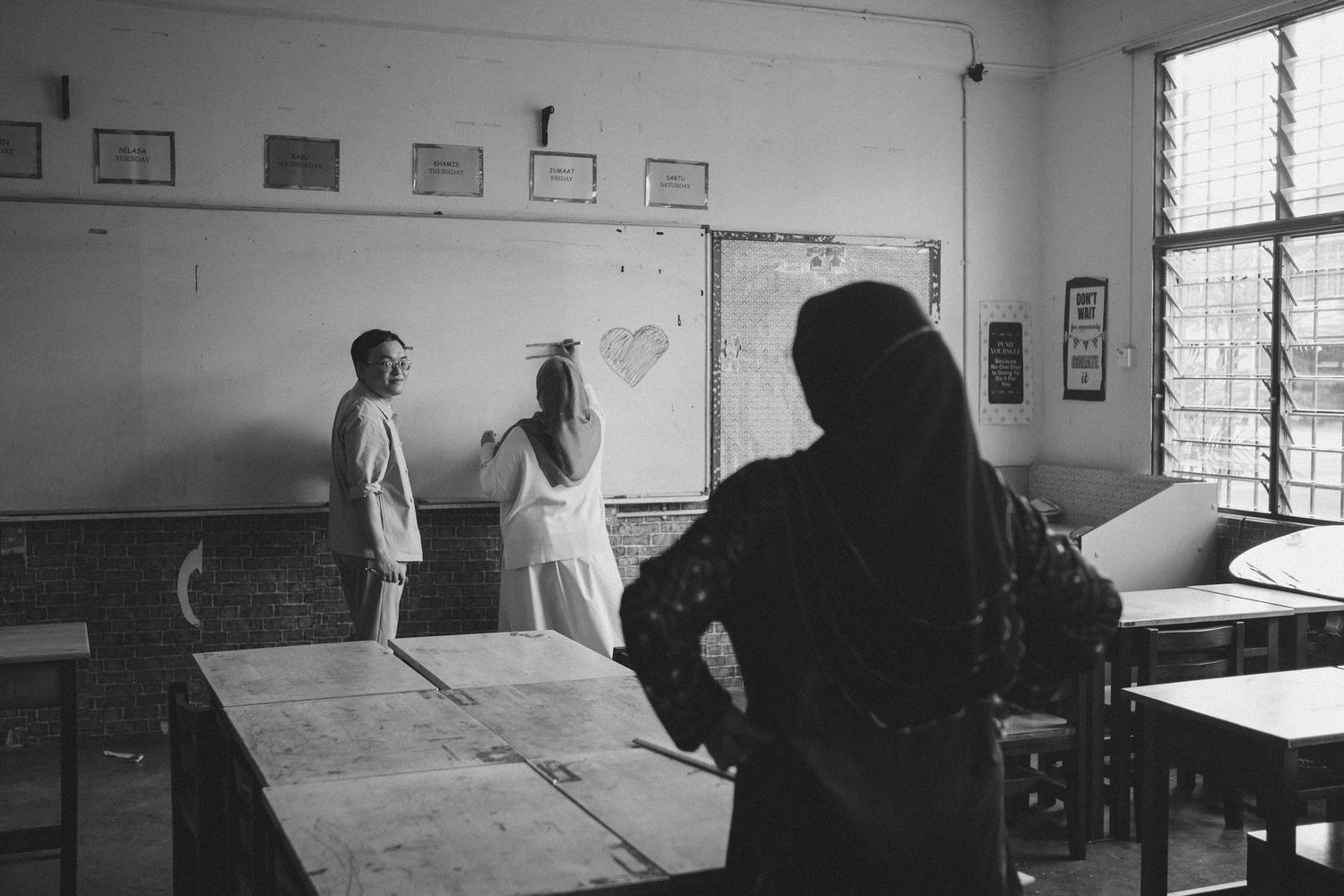 Black and white photo of students writing on chalkboard in empty classroom.