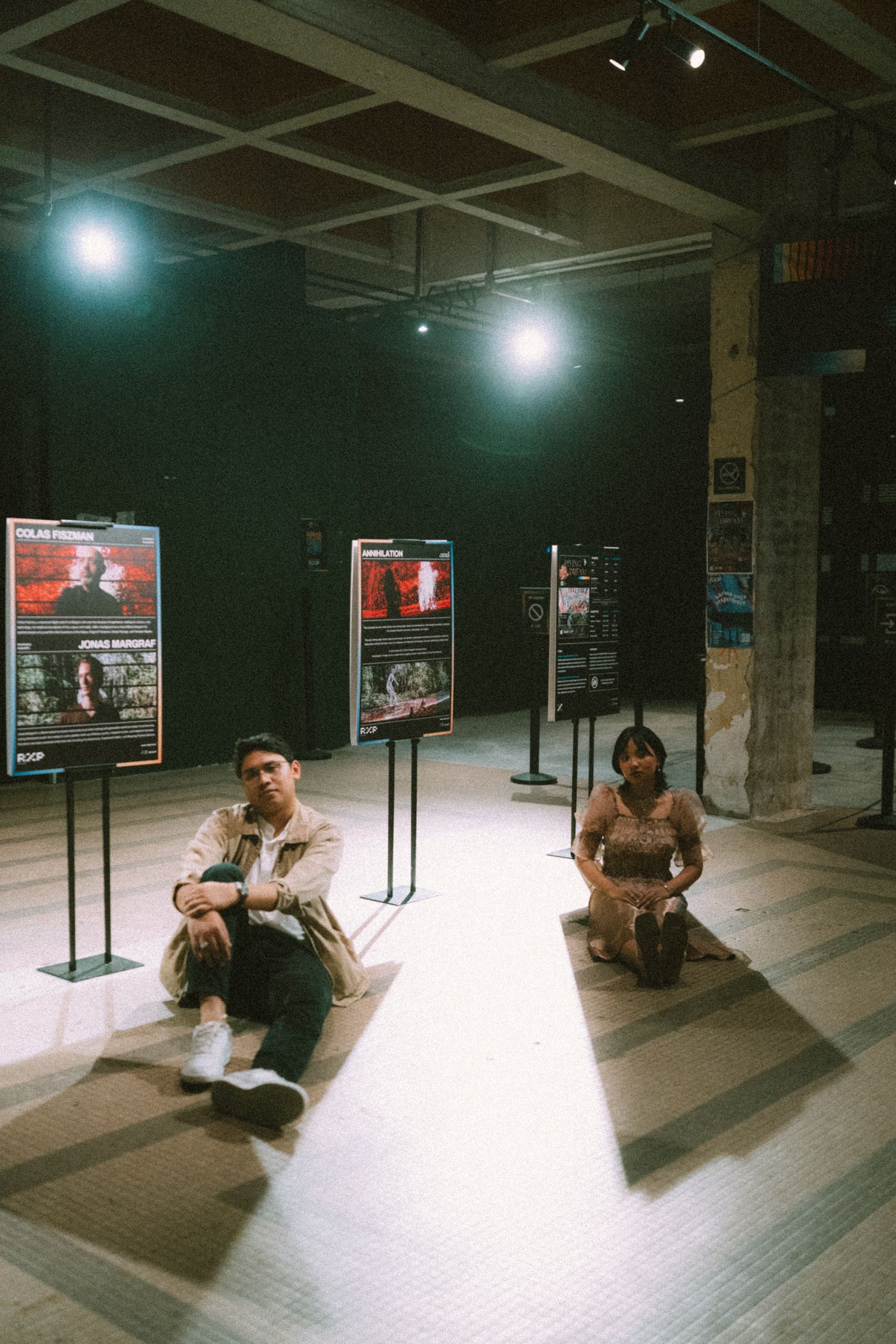Two people sit on a floor with movie posters displayed on stands in a dimly lit indoor space.