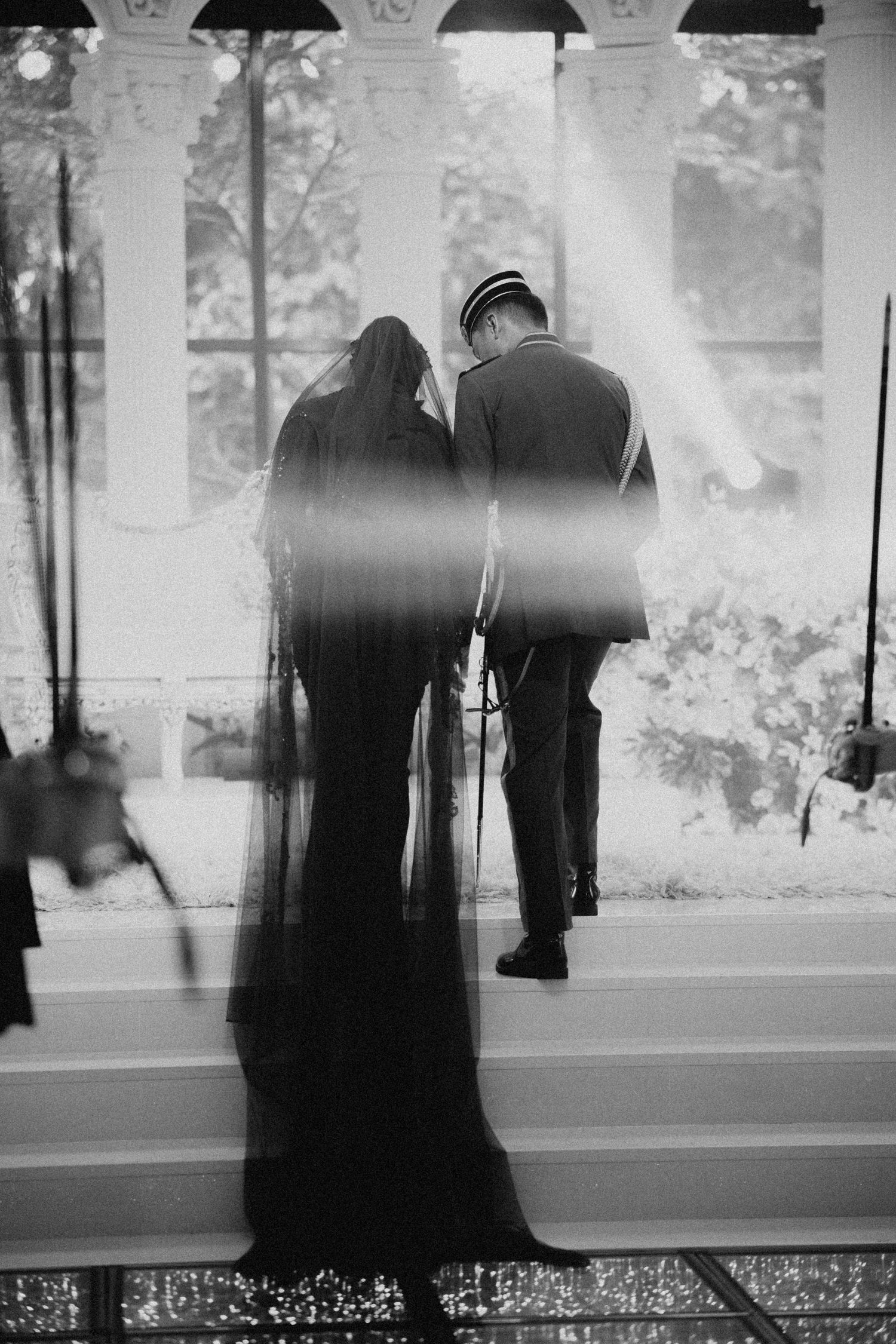 Black and white silhouette photograph of a couple standing on stairs between white columns in a romantic vintage scene.