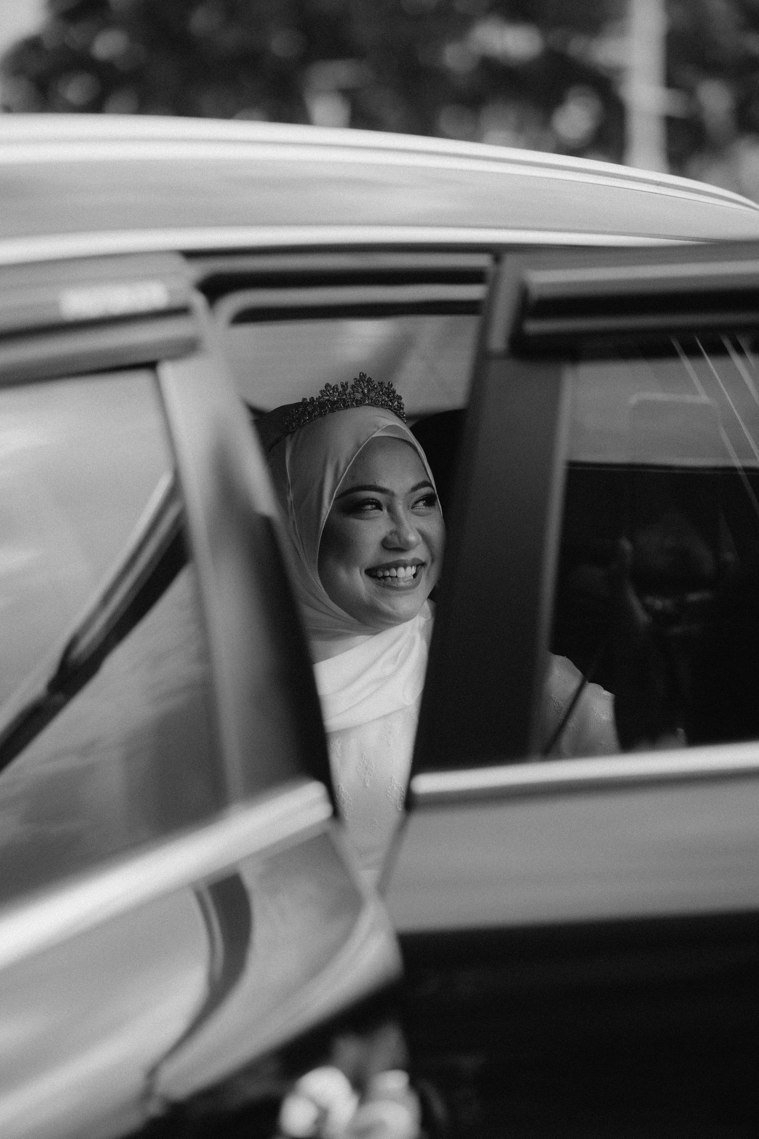 Black and white photograph of someone wearing a hijab and tiara smiling through a car window.